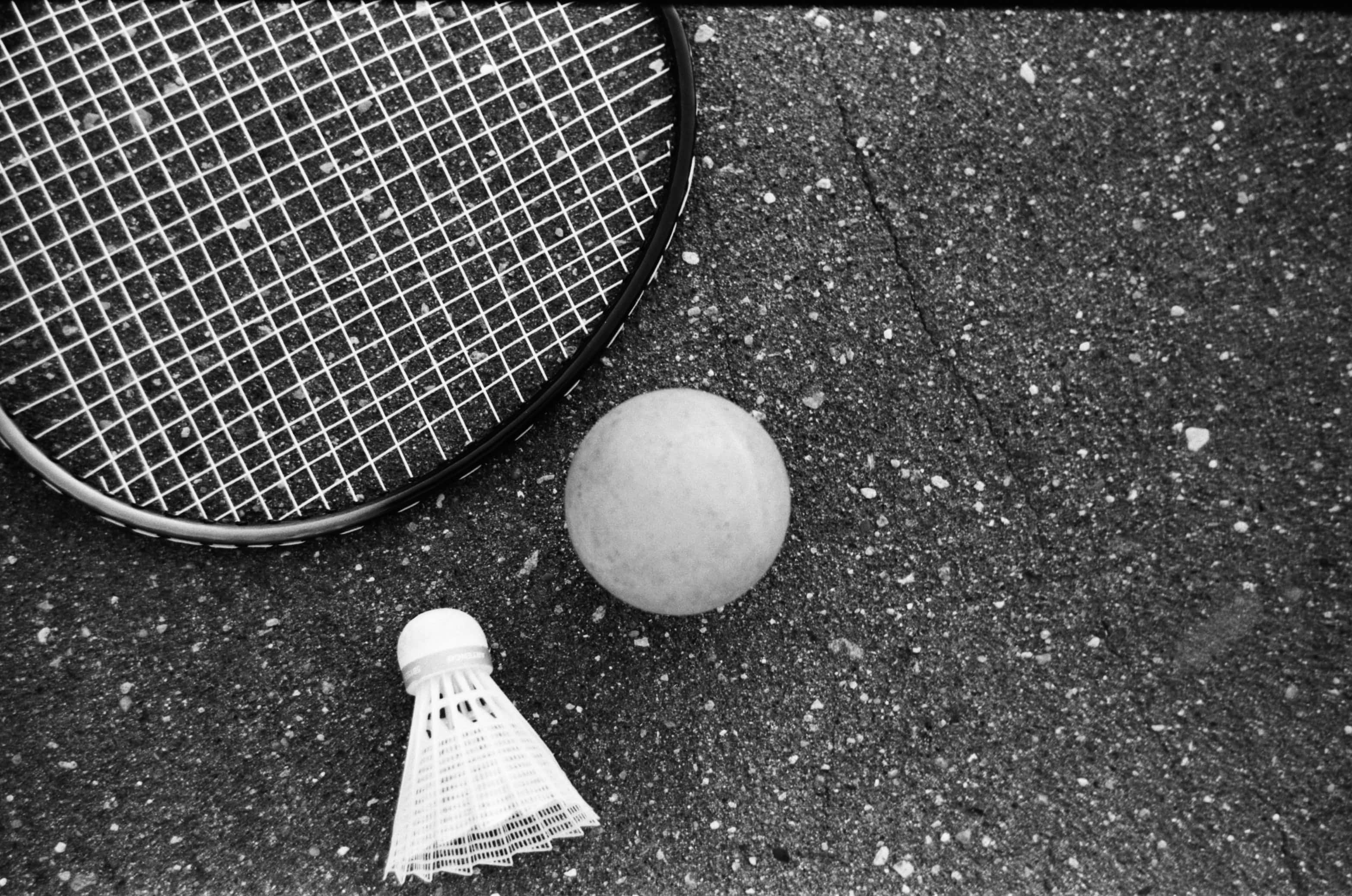 Badminton 桌布 壁紙 — 一個黑白特寫，顯示羽毛球拍、羽毛球和球放在破裂的瀝青上。