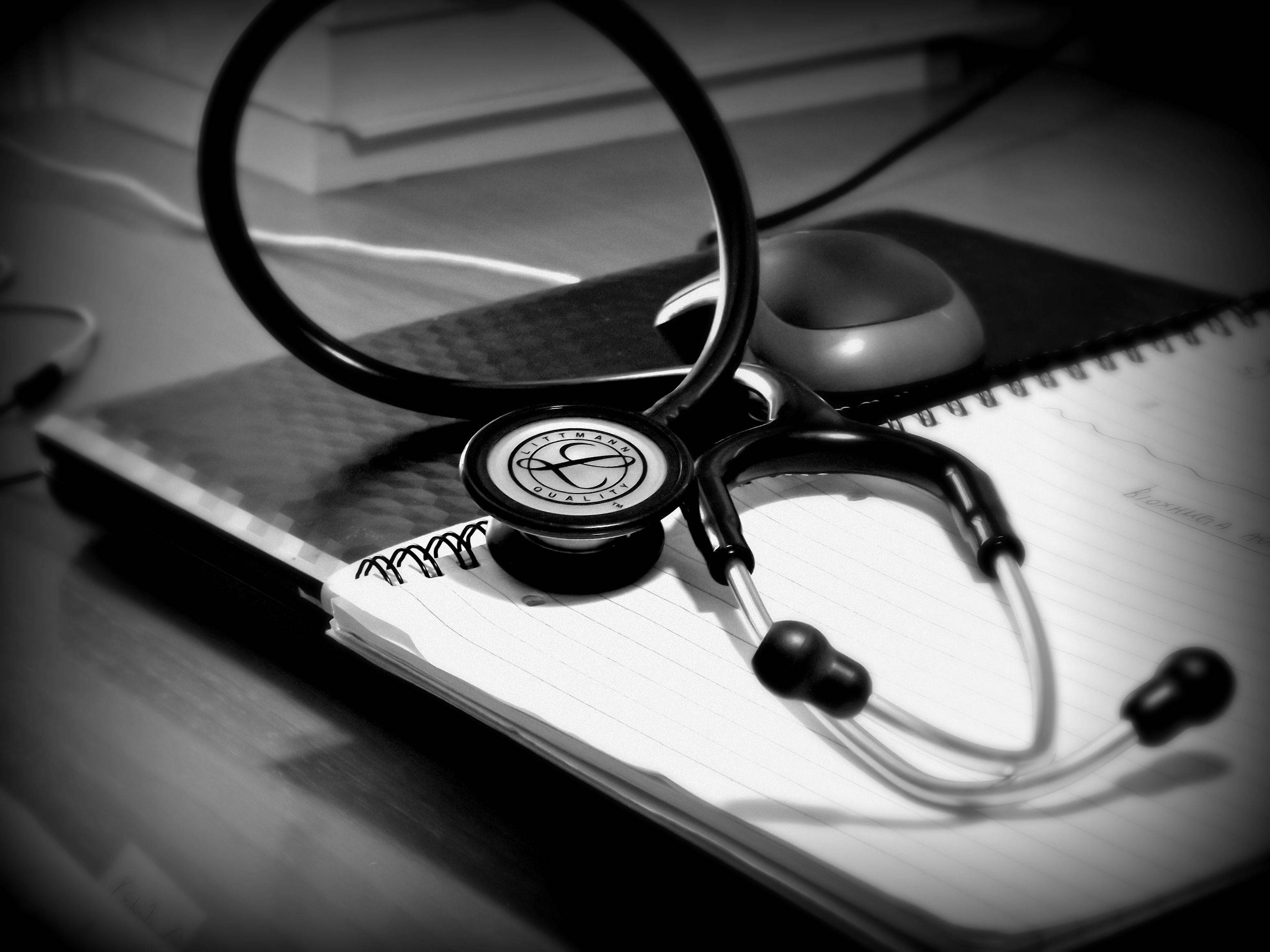 Stethoscope 桌布 壁紙 — 一個黑白聽診器放在木桌上打開的筆記本上，附近有一個電腦滑鼠。