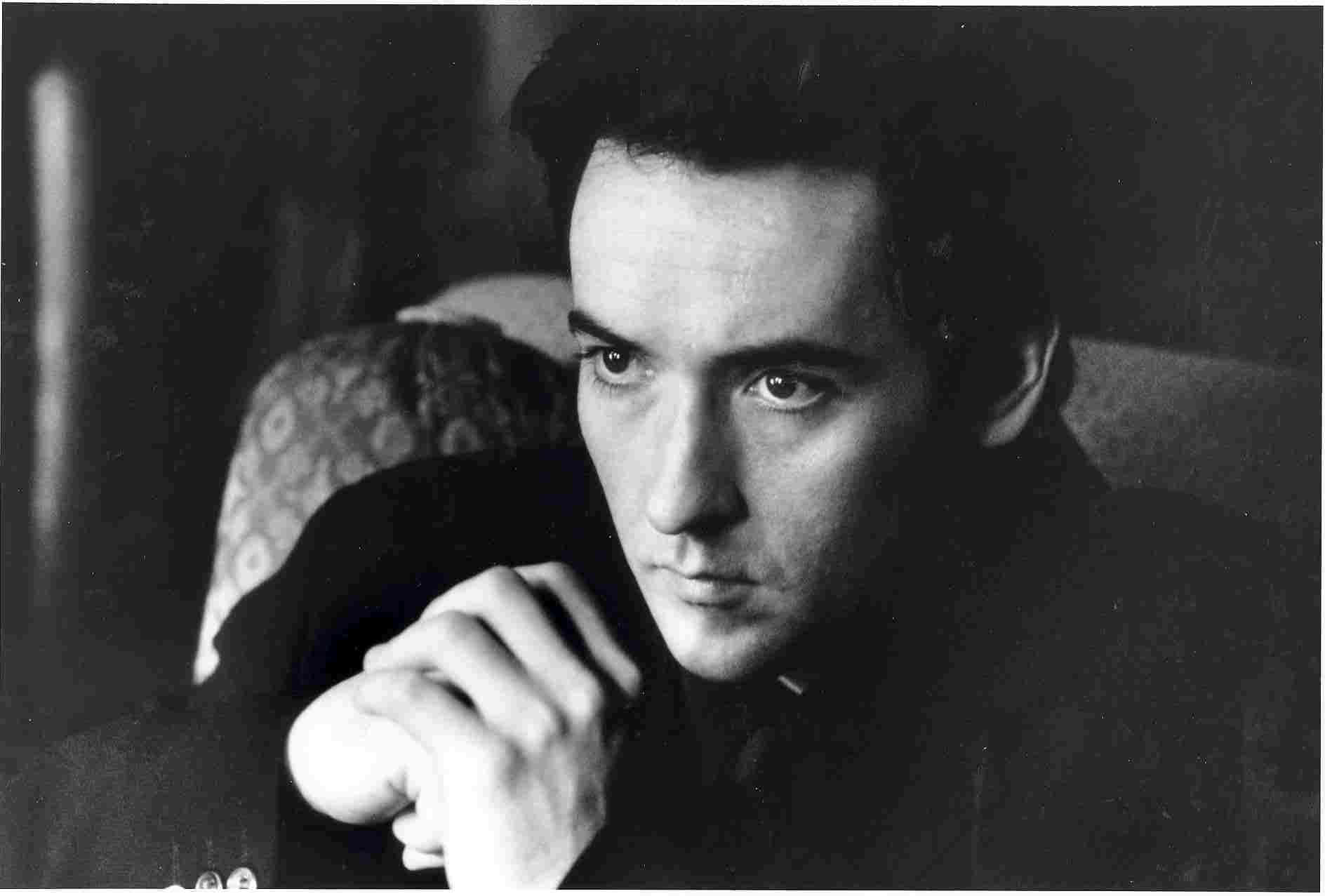 John Cusack 桌布 壁紙 — 一個黑白肖像，捕捉一位眼神銳利的男士，在昏暗的房間裡將下巴擱在交疊的雙手上。