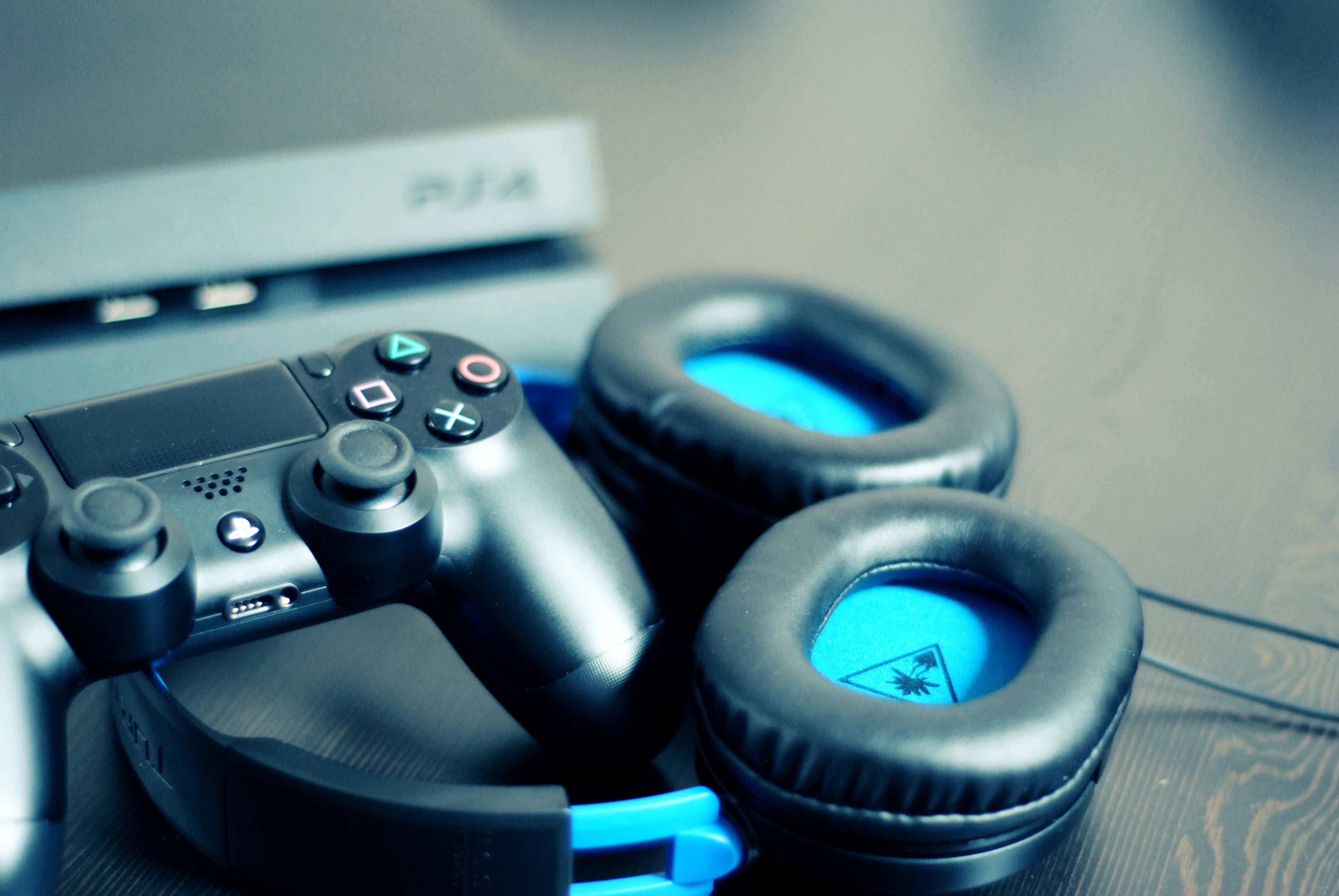 Gaming Consoles 桌布 壁紙 — 一個黑色PS4手掣同藍色發光耳機嘅特寫，放喺木質表面上，背景有模糊嘅主機。