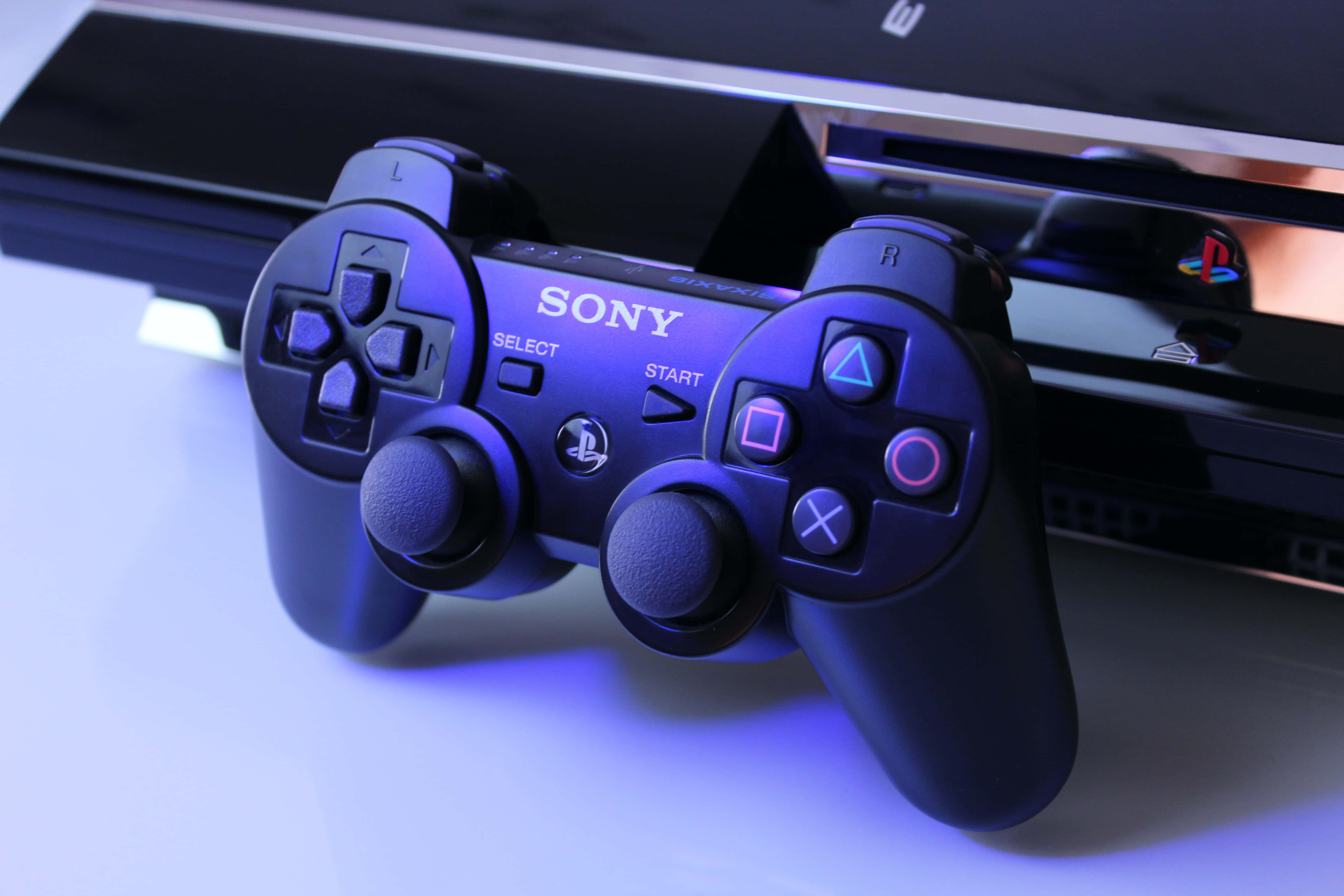 Gaming Consoles 桌布 壁紙 — 一個黑色Sony PlayStation 4手掣喺藍光下發光，旁邊係一個白色表面上嘅主機。