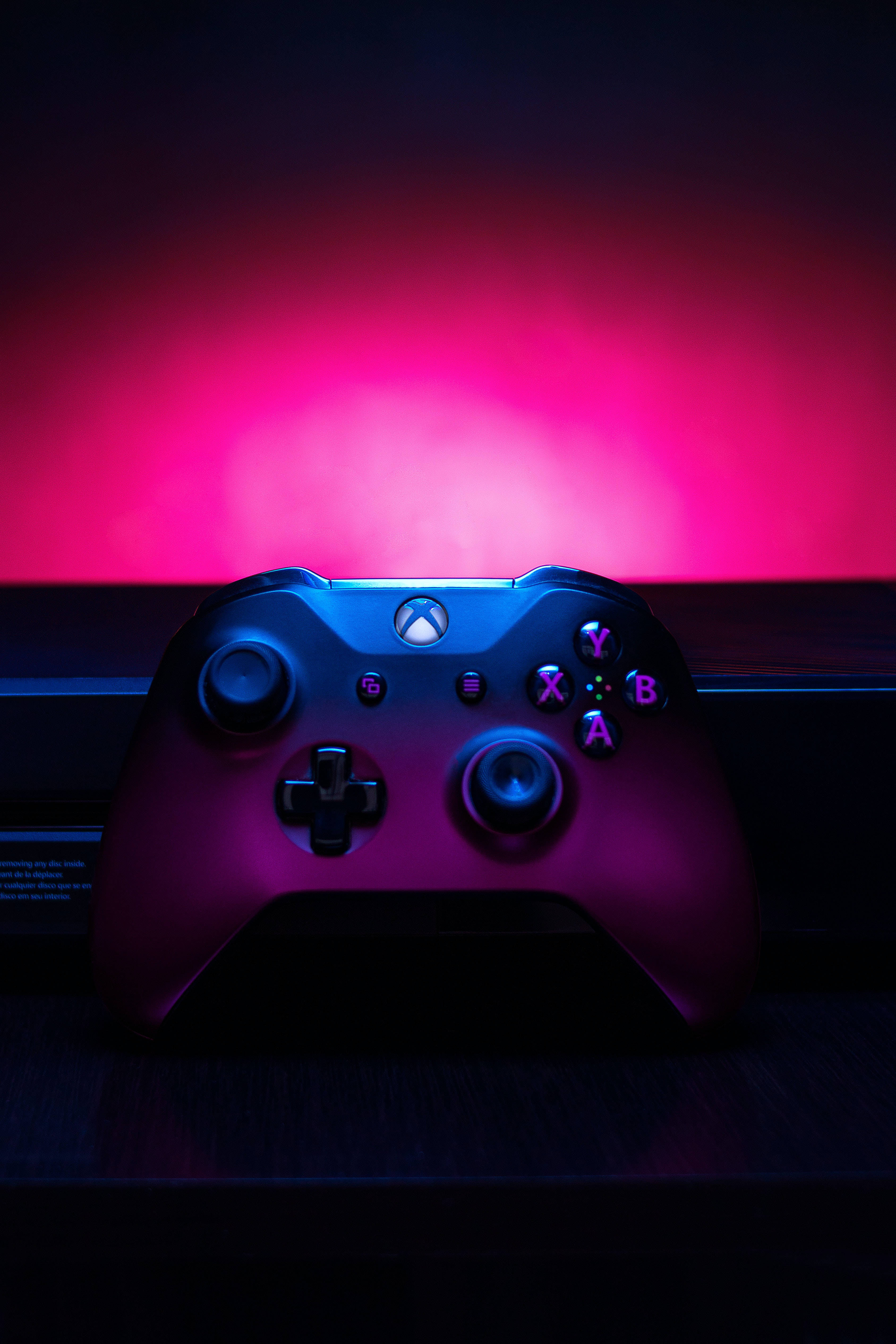 Dark Purple iPhone 桌布 壁紙 — 一個黑色Xbox手掣在深色背景上沐浴在充滿活力的粉紅和藍色霓虹燈光中。