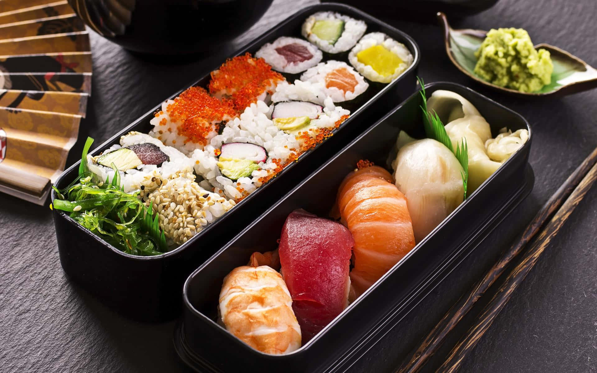 Sushi 桌布 壁紙 — 一個黑色便當盒裝滿色彩繽紛的壽司卷和刺身，整齊地排列在深色石板表面上，配以芥末和筷子。