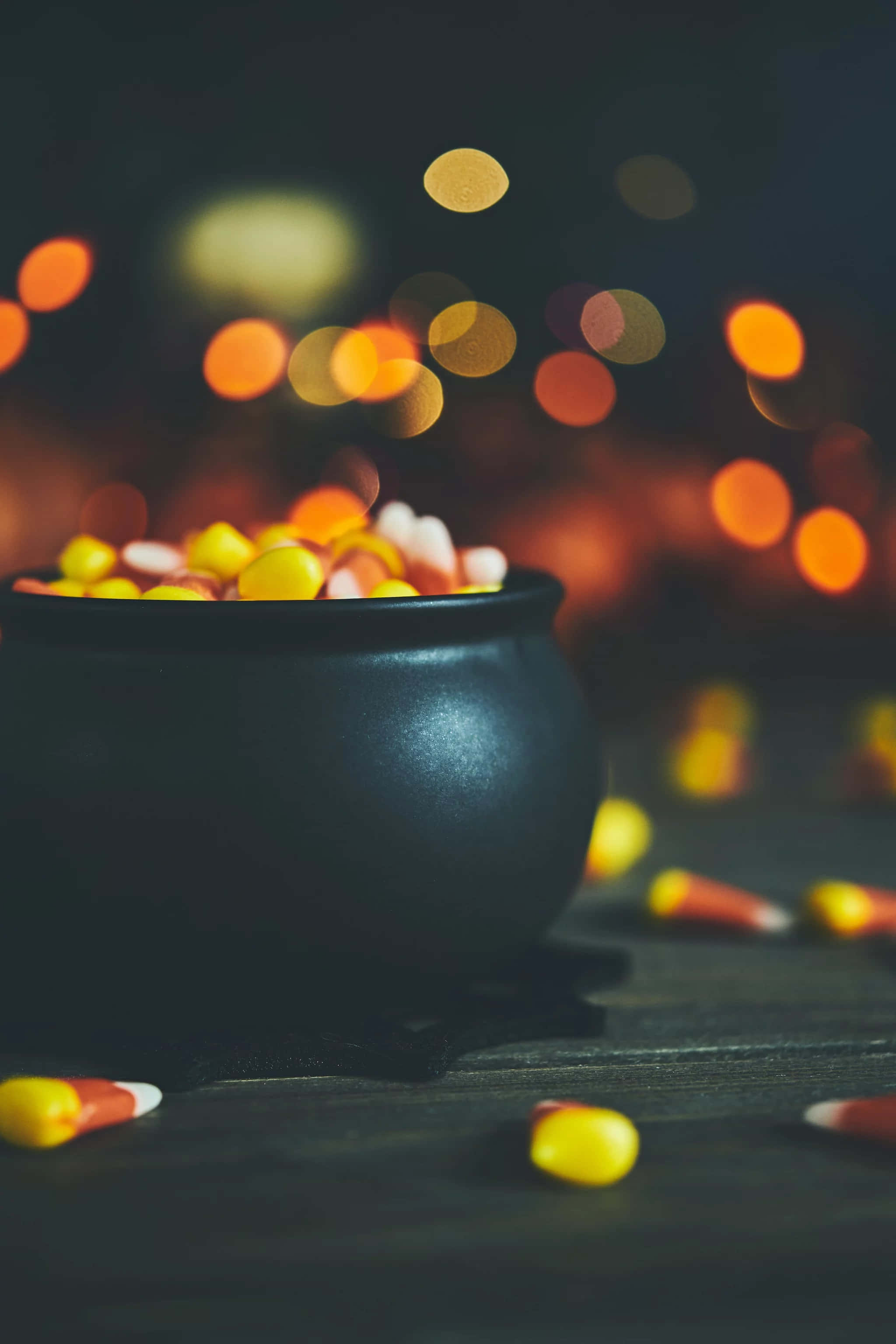 Halloween Phone 桌布 壁紙 — 一個黑色大鍋裝滿了糖果粟米，放在深色木桌上，背景有溫暖的散景燈光。