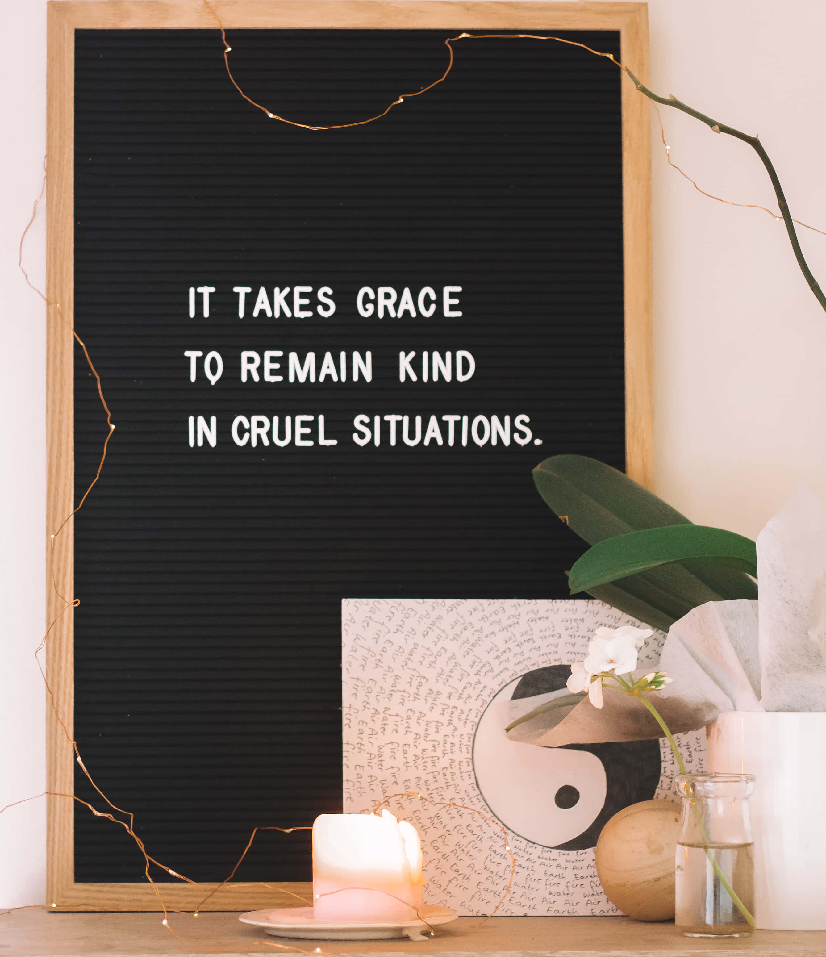 Soulful 桌布 壁紙 — 一個黑色字母板，上面有白色文字寫著「IT TAKES GRACE TO REMAIN KIND IN CRUEL SITUATIONS」，周圍環繞著點燃的蠟燭、綠色植物和串燈，放在木架上。