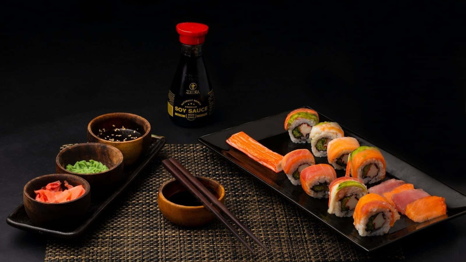 Sushi 桌布 壁紙 — 一個黑色托盤上放著色彩繽紛的壽司卷和刺身，旁邊有醬油、生薑、芥末和筷子，置於深色編織墊上。