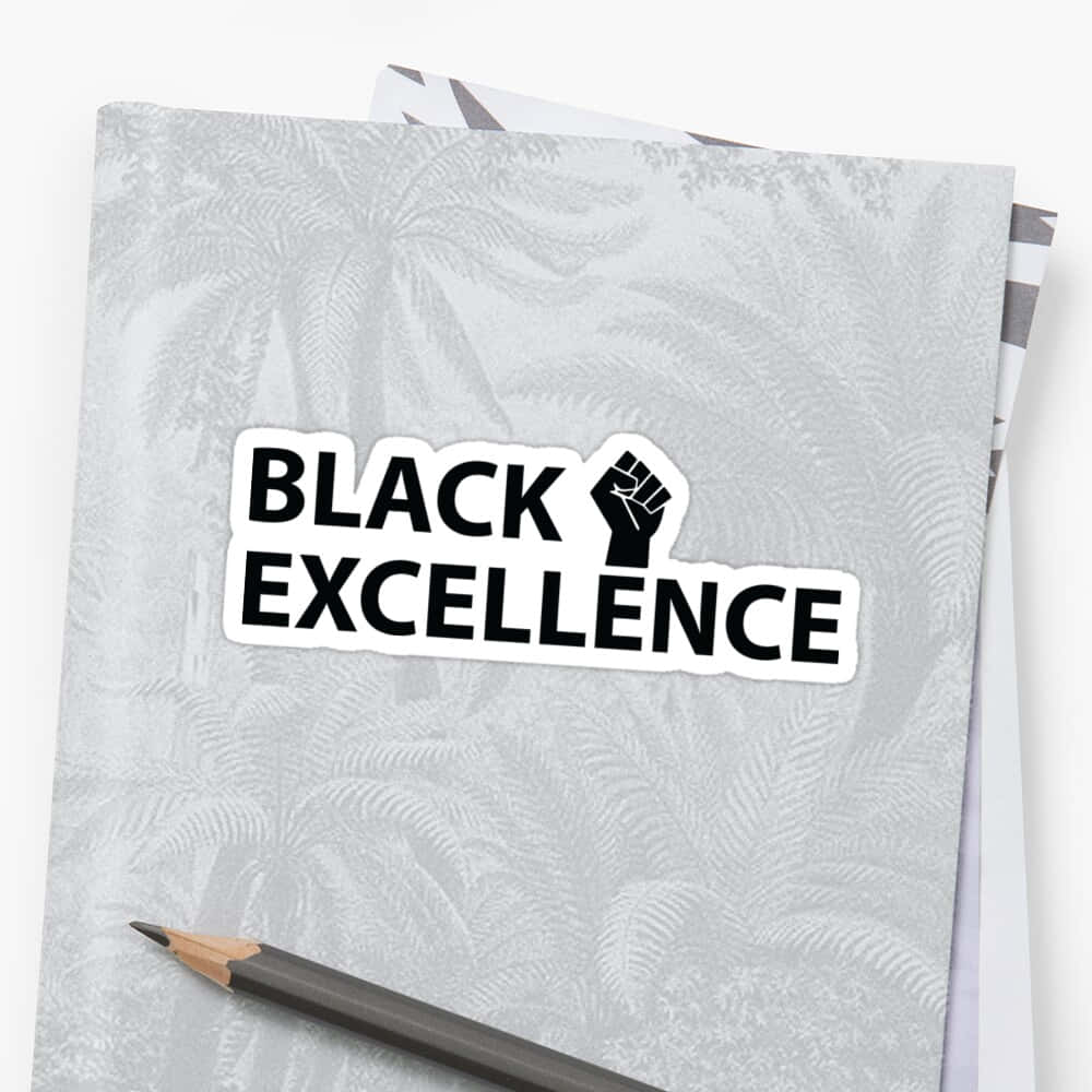 Black Excellence 桌布 壁紙 — 一個黑色拳頭貼紙，上面有粗體字「BLACK EXCELLENCE」，放在灰色蕨類圖案筆記本上，旁邊有一支鉛筆。