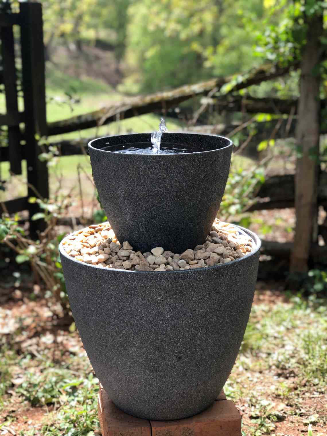 戶外 桌布 壁紙 — 一個黑色斑點兩層戶外噴泉，水從頂部到底部層層流下，周圍環繞著鵝卵石和綠色植物。