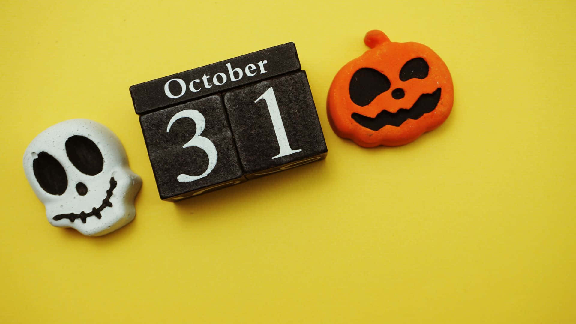 October 31st 桌布 壁紙 — 一個黑色木製日曆塊顯示10月31日，兩側是白色骷髏頭和橙色南瓜，背景為亮黃色。