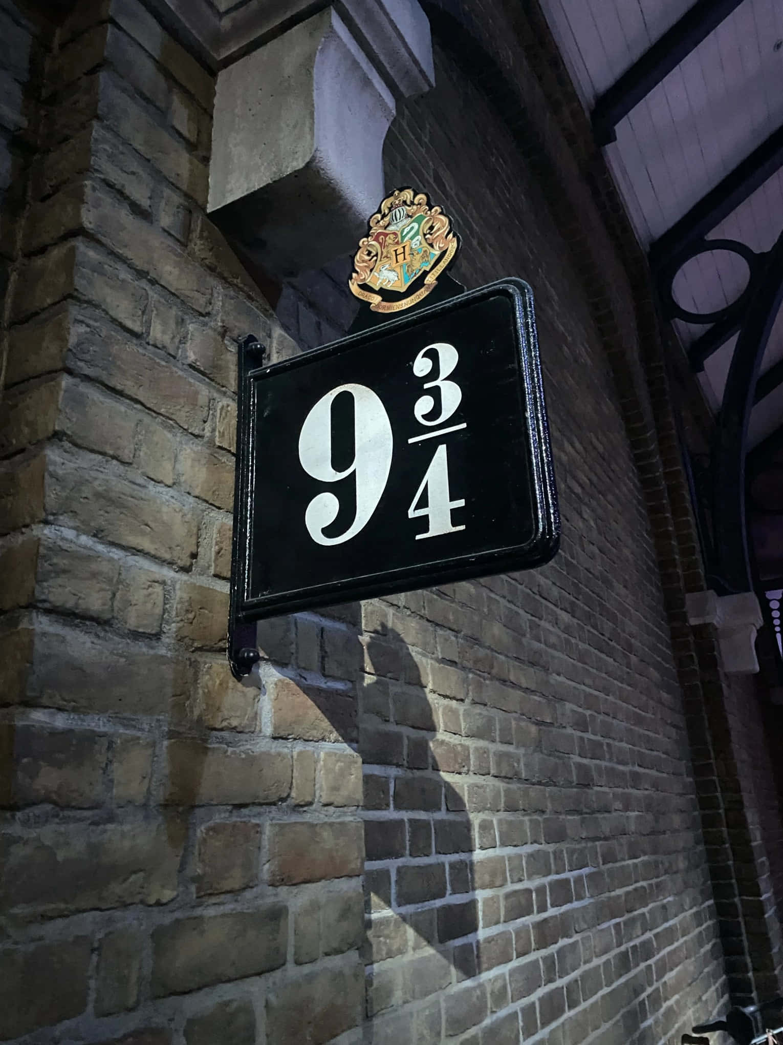 Platform 9 3/4 桌布 壁紙 — 一個黑色標誌顯示9 3/4，帶有霍格華茲徽章，安裝在磚牆上，光線昏暗。