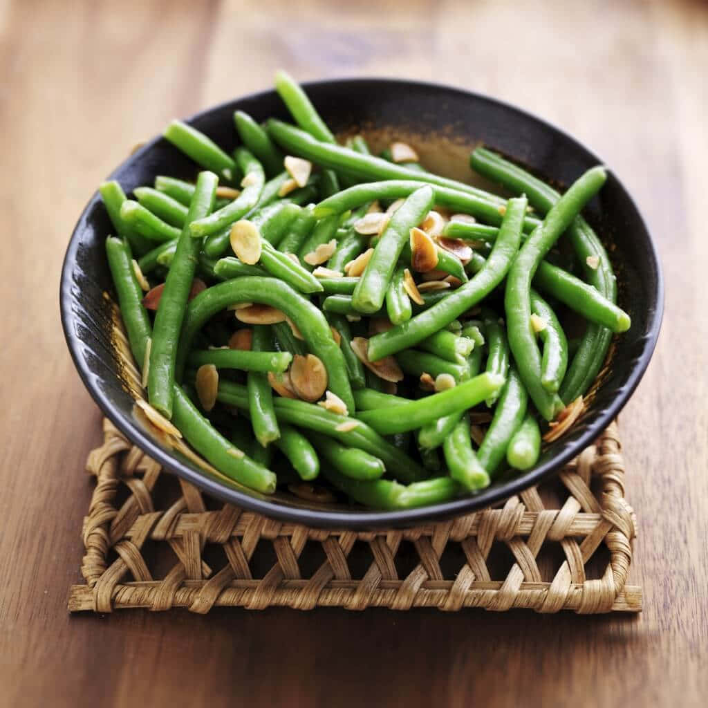 beans 桌布 壁紙 — 一個黑色碗裝滿鮮豔的四季豆，上面放有烤杏仁片，放在木桌上的編織墊上。