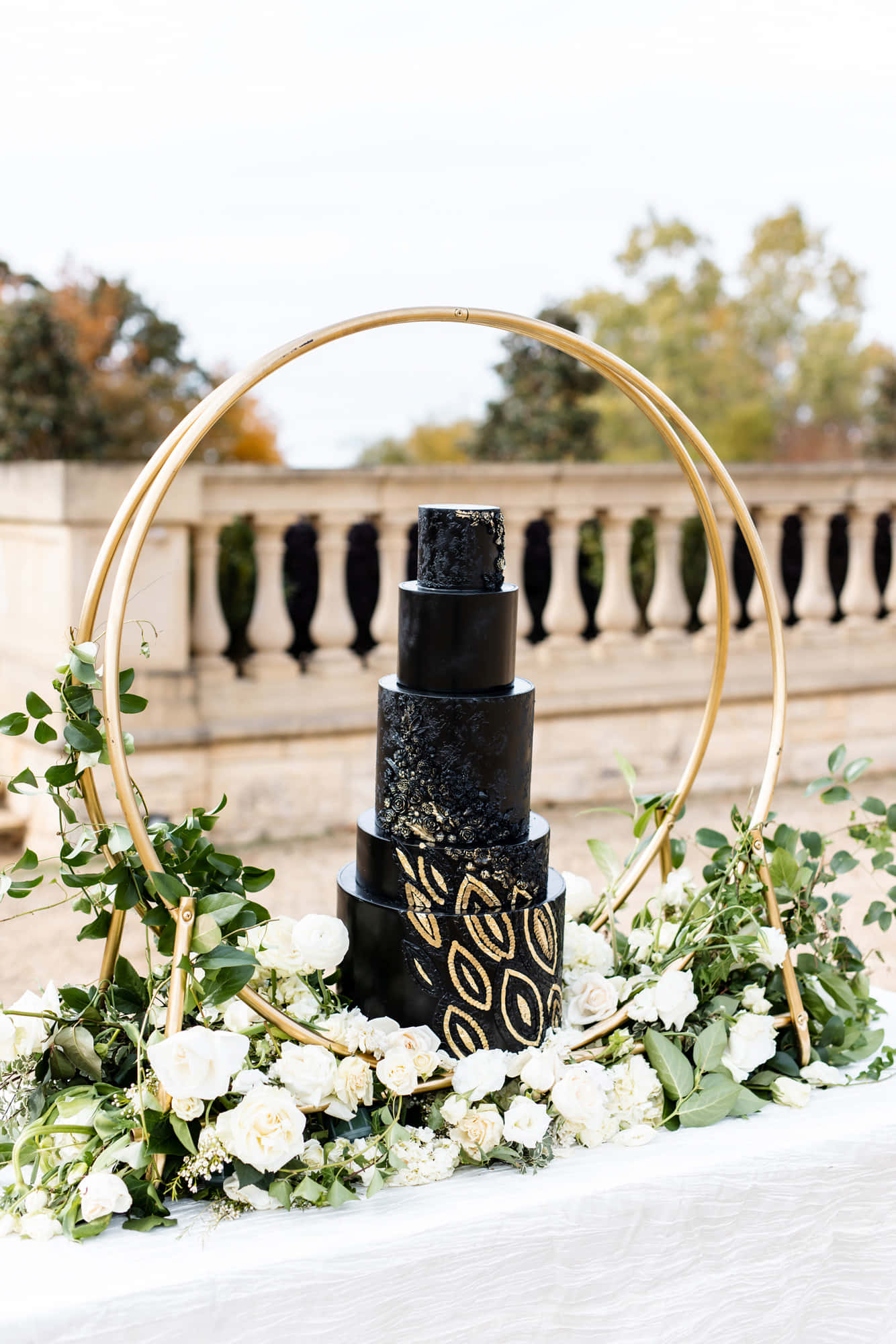 Black And White Wedding 桌布 壁紙 — 一個黑色紋理婚禮蛋糕，帶有金箔圖案，放在一個金色圓環下，周圍環繞著白玫瑰和綠色植物。