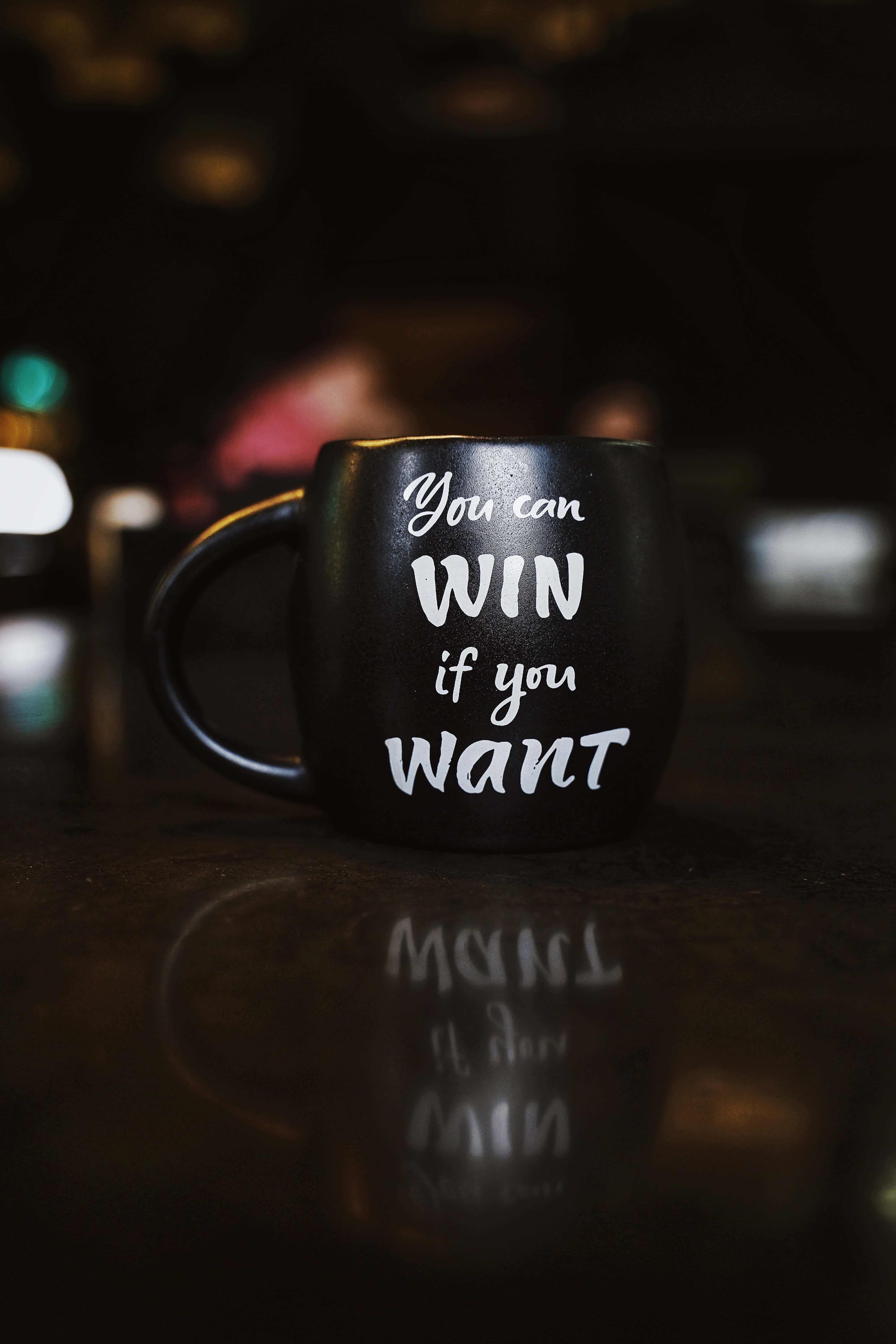Success 桌布 壁紙 — 一個黑色馬克杯帶有白色勵志文字，放置在昏暗光線下的反光表面上。