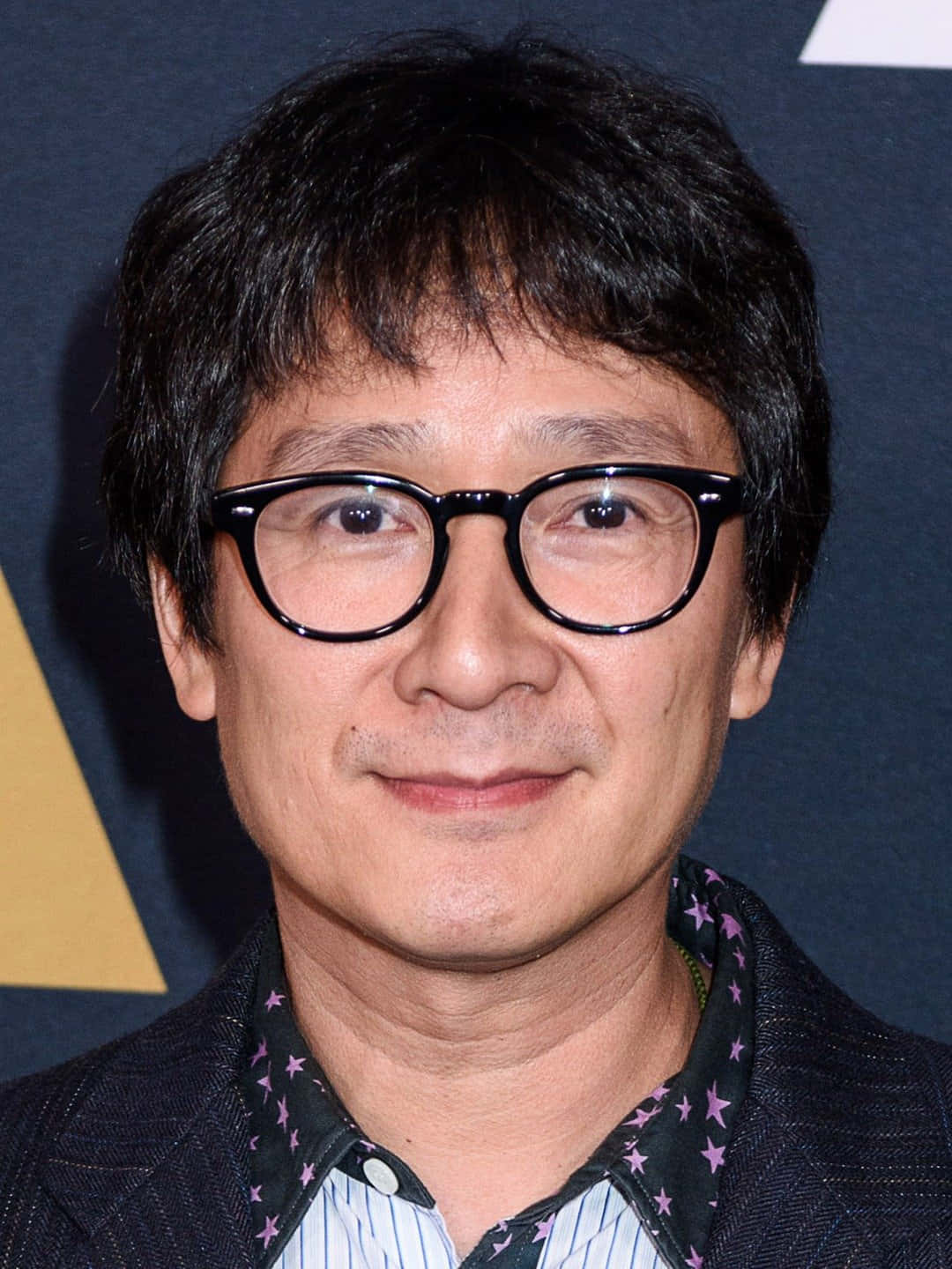 Ke Huy Quan 桌布 壁紙 — 一個黑髮、戴粗框眼鏡的男人近距離肖像，對著鏡頭溫柔微笑。