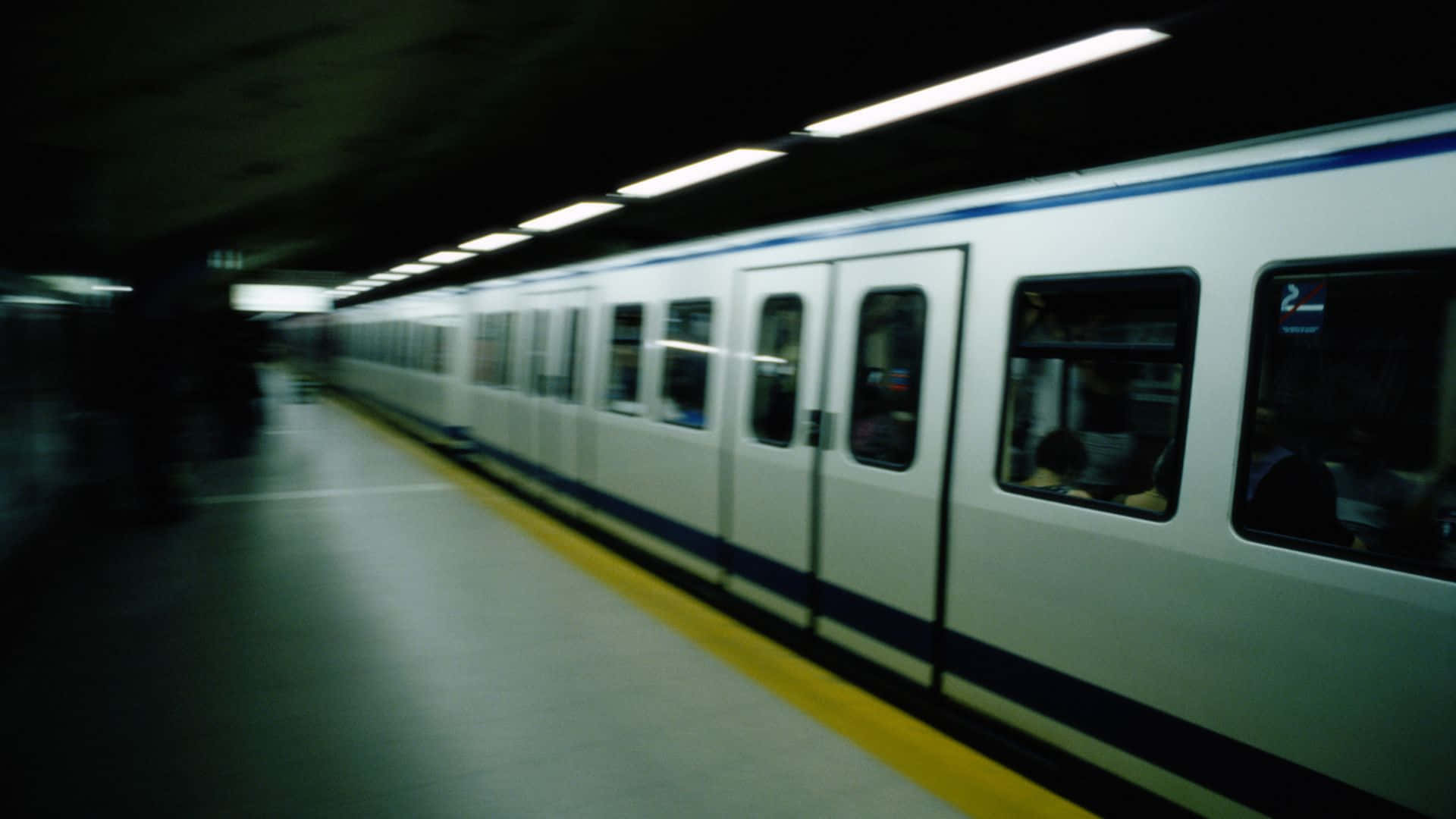 Subway 桌布 壁紙 — 一列模糊的地鐵列車高速駛過一個燈光昏暗的月台，月台上有黃色安全線和頭頂燈光。