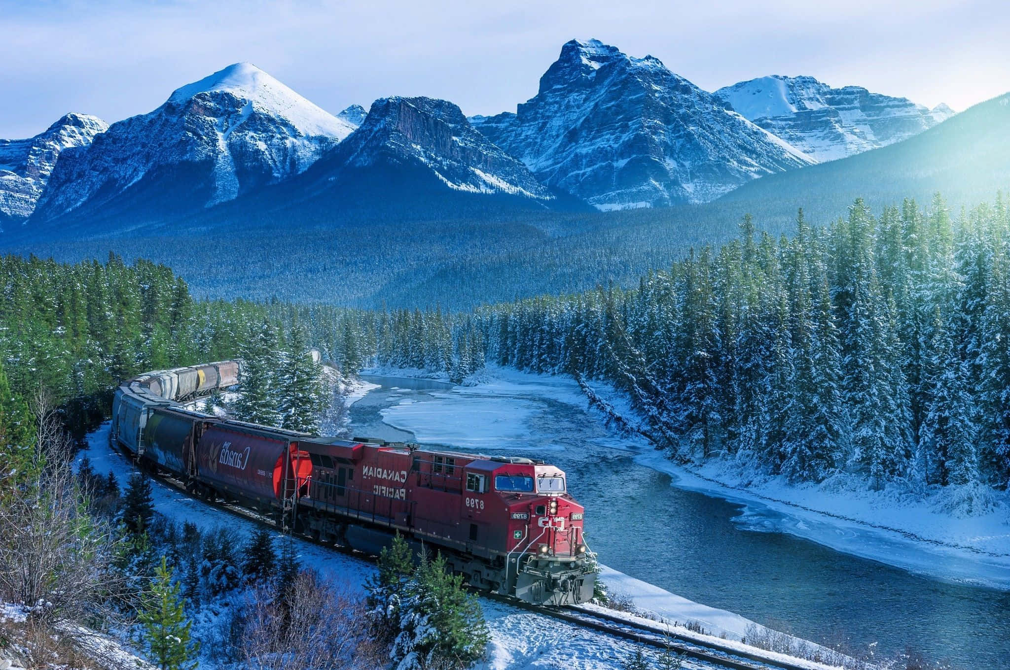 Canada 桌布 壁紙 — 一列紅色的加拿大太平洋鐵路火車沿著結冰河流旁的積雪軌道蜿蜒而行，四周是高聳的雪山和常綠森林，上方是明亮的藍天。