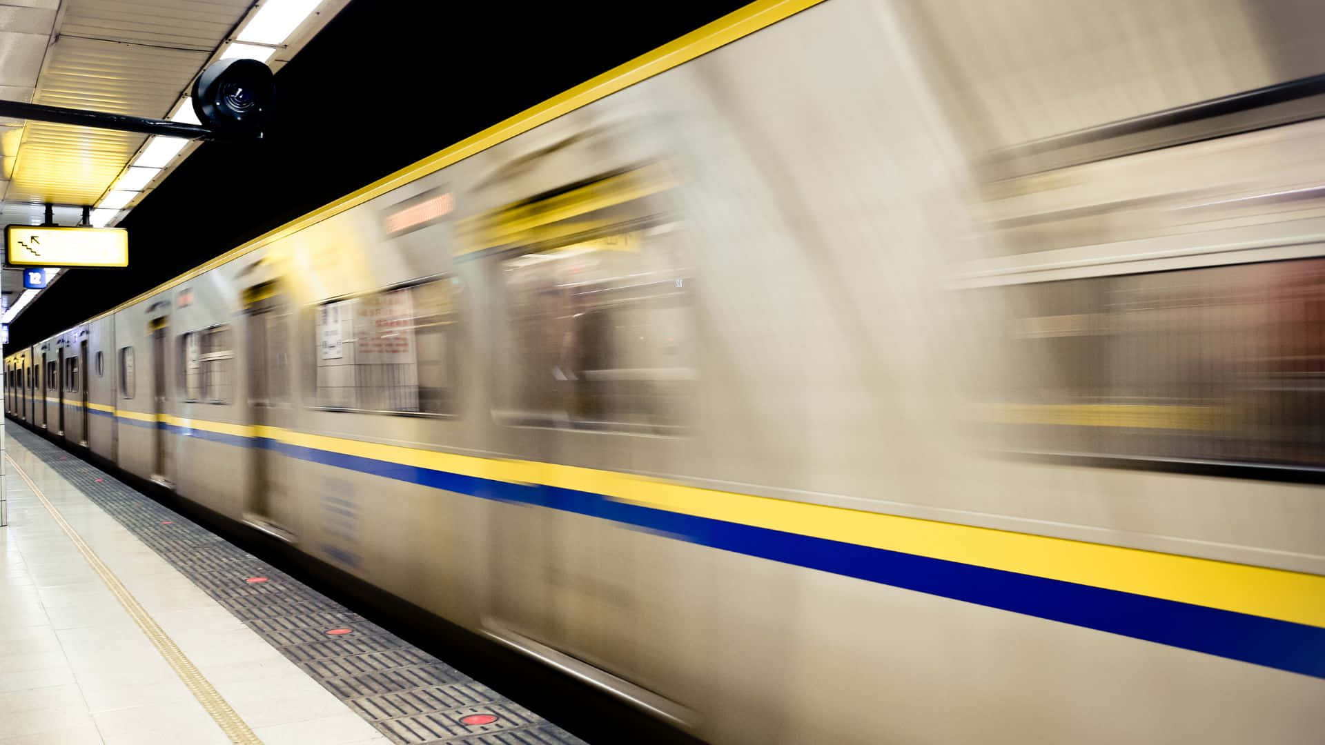 Subway 桌布 壁紙 — 一列銀色地鐵列車帶有黃色和藍色條紋，在明亮的燈光下高速駛過車站月台。