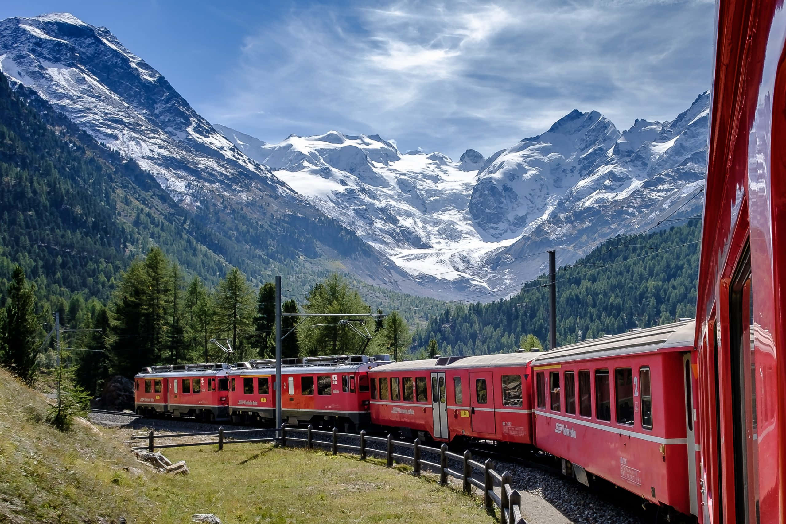 Switzerland 桌布 壁紙 — 一列鮮紅色的火車蜿蜒穿過一個鬱鬱蔥蔥的綠色山谷，兩旁是高聳的雪山，上方是明亮的藍天。
