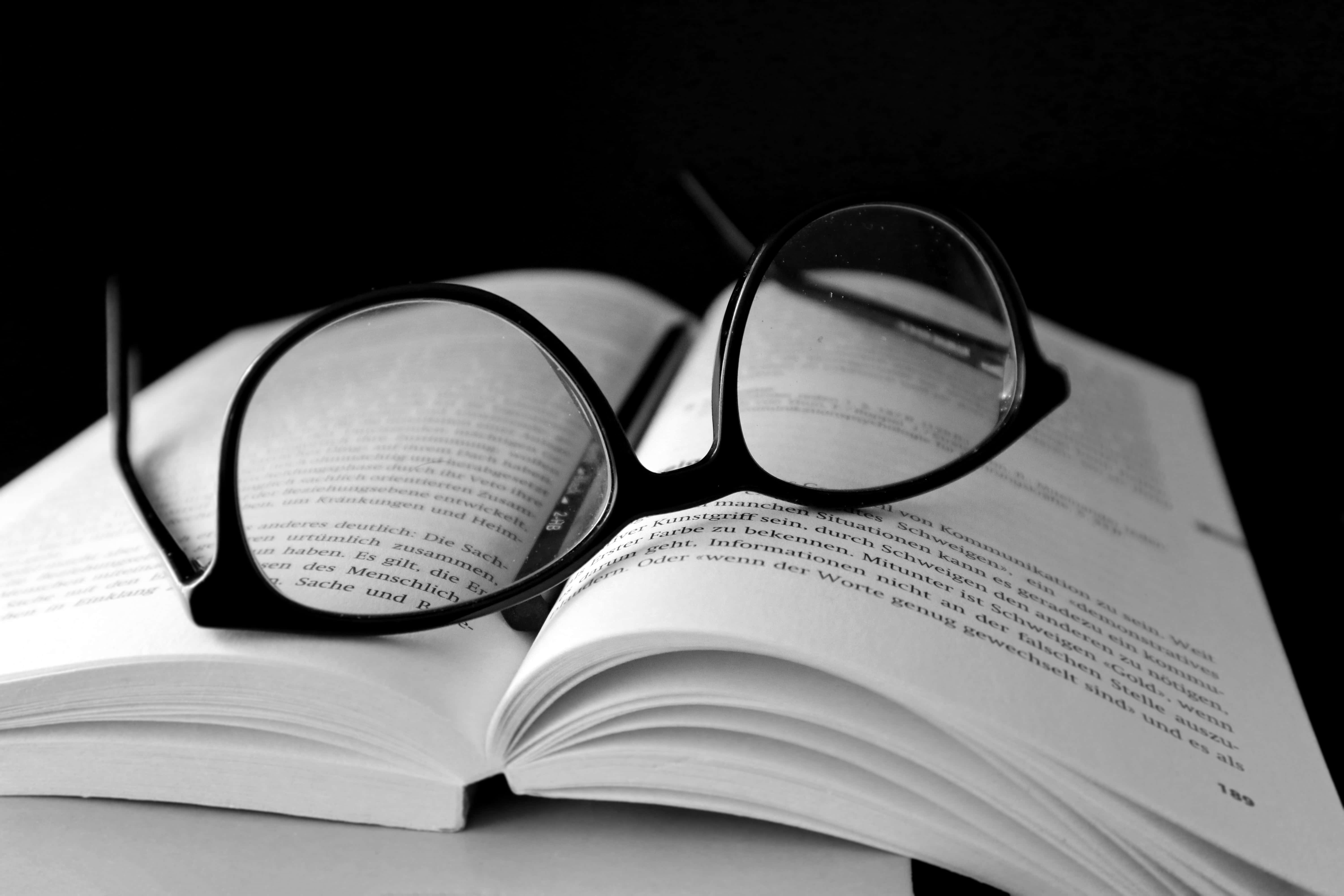 Black And White Book 桌布 壁紙 — 一副黑框眼鏡擱在一本打開的書上，書頁上的文字清晰可見，背景為純黑色。