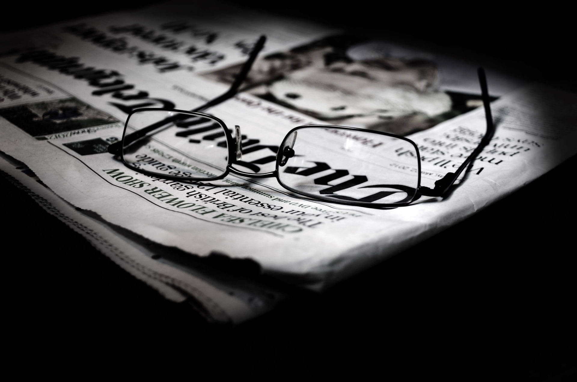 Newspaper 桌布 壁紙 — 一副黑框眼鏡放在一堆報紙上，處於黑暗房間中的戲劇性聚光燈下。