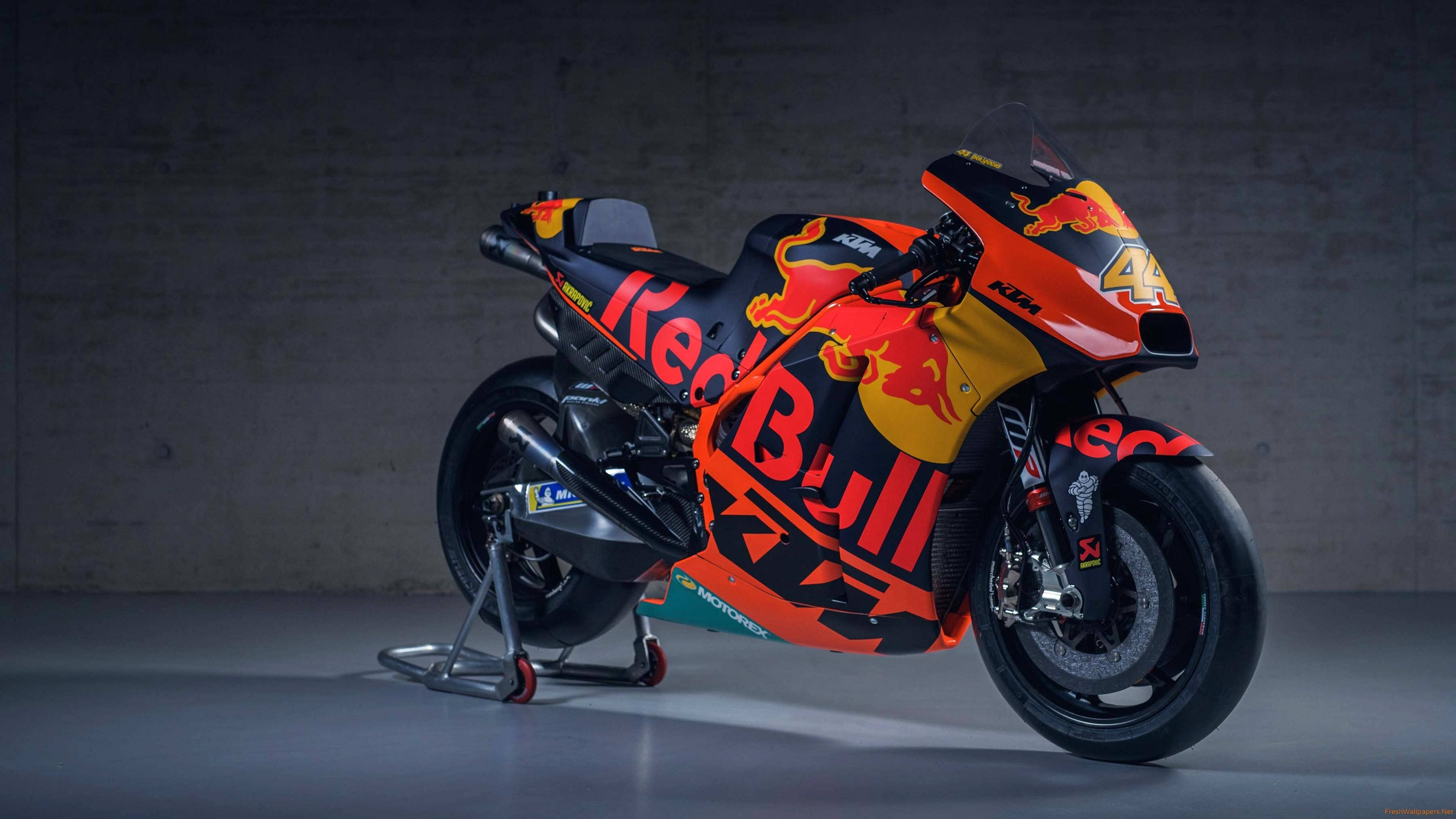 機車 桌布 壁紙 — 一台時尚的Red Bull KTM MotoGP賽車，帶有鮮豔的橙色和黑色塗裝，停在支架上，背景是紋理混凝土牆。