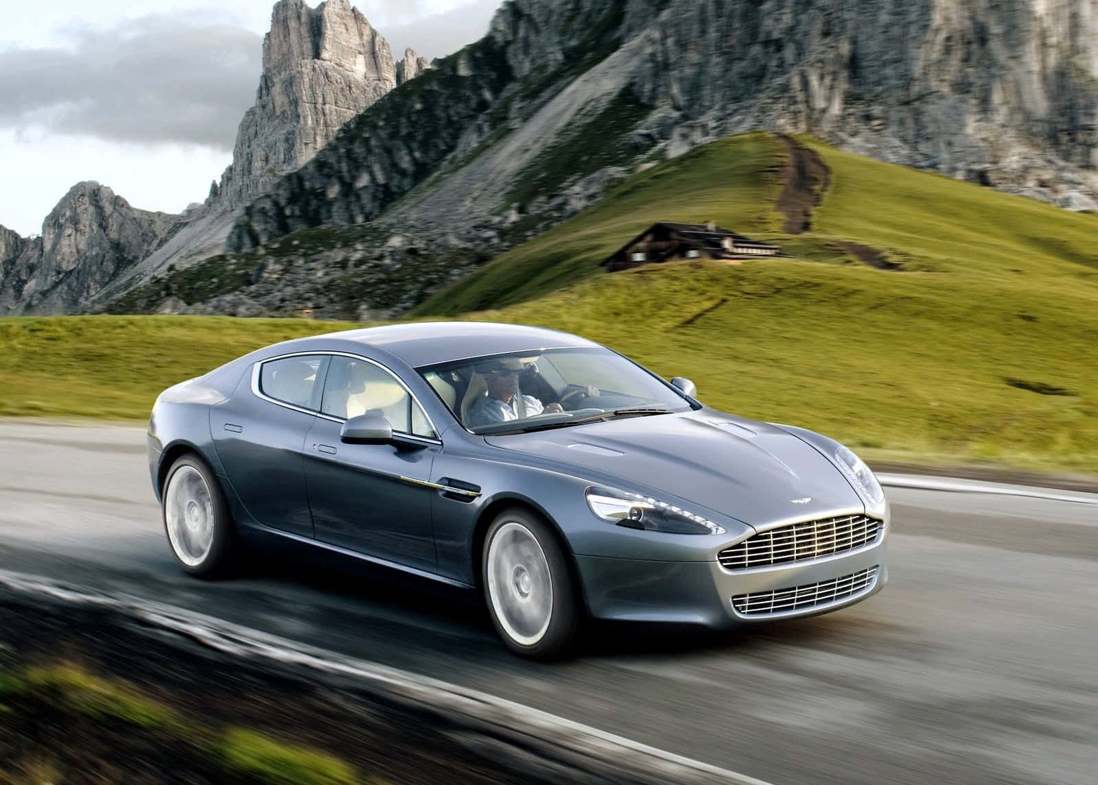 Aston Martin Rapide S 桌布 壁紙 — 一台時尚的銀色阿斯頓馬丁沿著蜿蜒的山路疾馳，背景是崎嶇的山峰和綠色的山丘。
