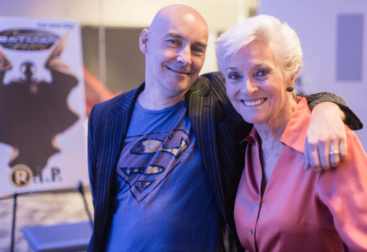 Grant Morrison 桌布 壁紙 — 一名光頭男子穿著藍色超人T恤和細條紋外套，在會議上微笑著擁抱一位穿著粉紅色襯衫的年長女士。