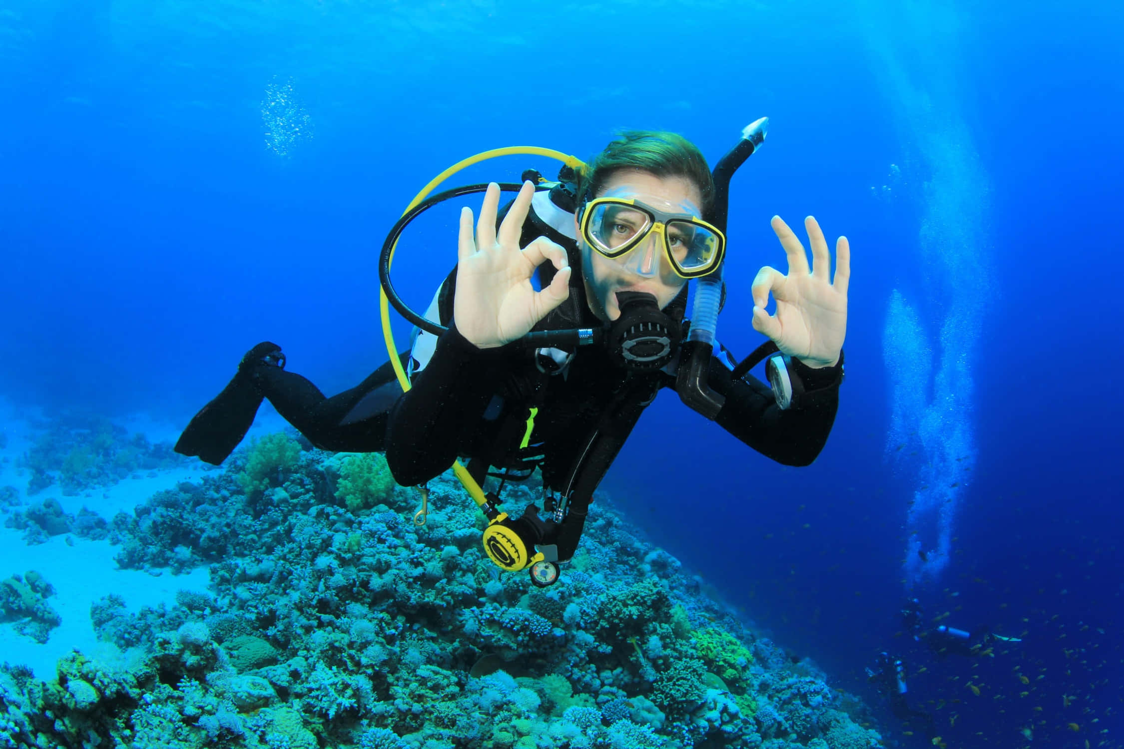 diving 桌布 壁紙 — 一名全副裝備的水肺潛水員在清澈藍色海水中的 vibrant 珊瑚礁上方游泳，豎起兩個大拇指。