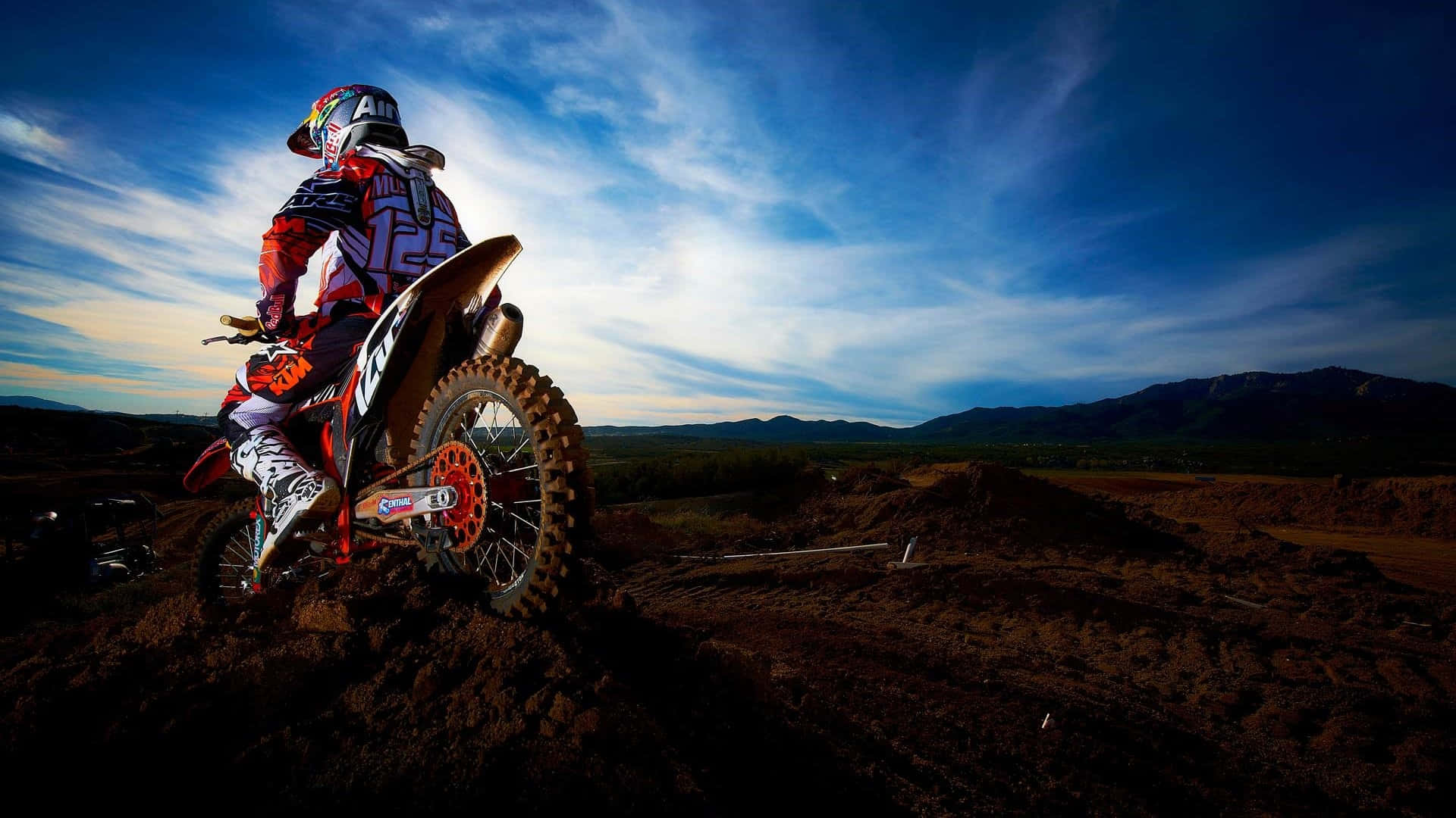 Motocross 桌布 壁紙 — 一名全副裝備的越野電單車手在泥丘上稍作休息，背景是戲劇性的暮色天空和遠山。