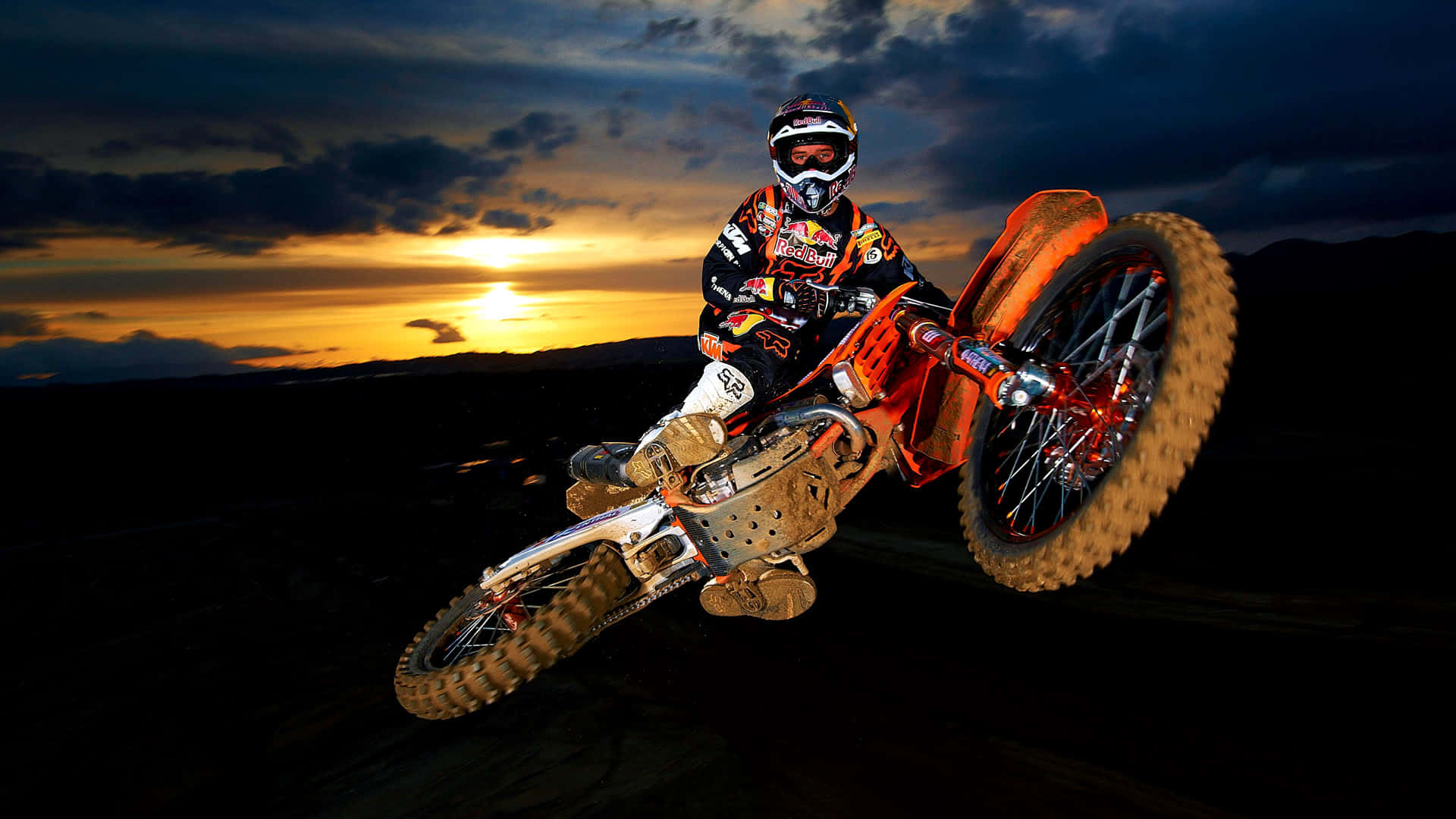 Motocross 桌布 壁紙 — 一名全副裝備的越野電單車手在金色雲彩的戲劇性日落天空下表演大膽的後輪平衡特技。