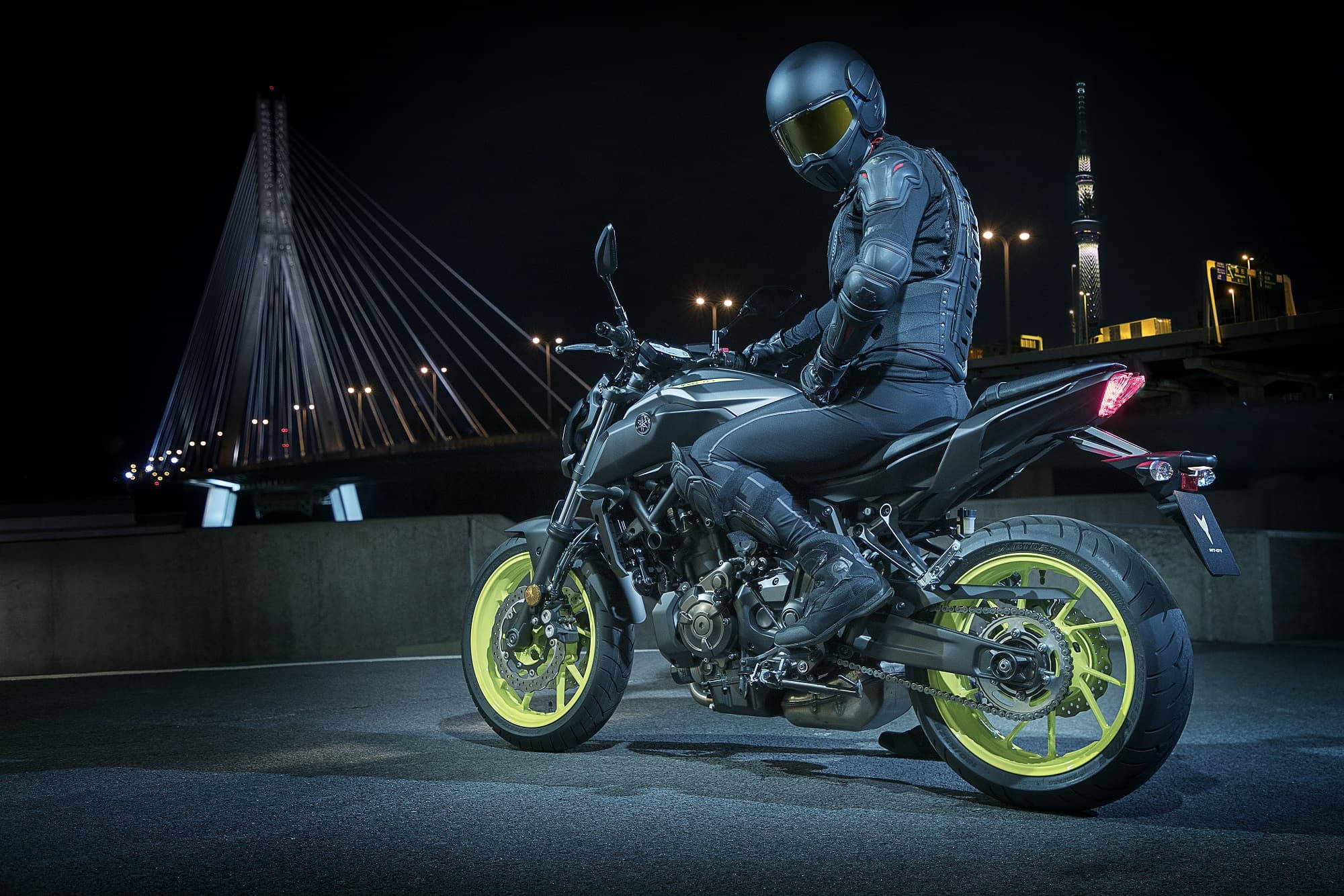yamaha mt 15 桌布 壁紙 — 一名全副裝備的騎士在夜間城市燈光下，坐在一輛配有螢光黃色車輪的時尚黑色電單車上，蓄勢待發。