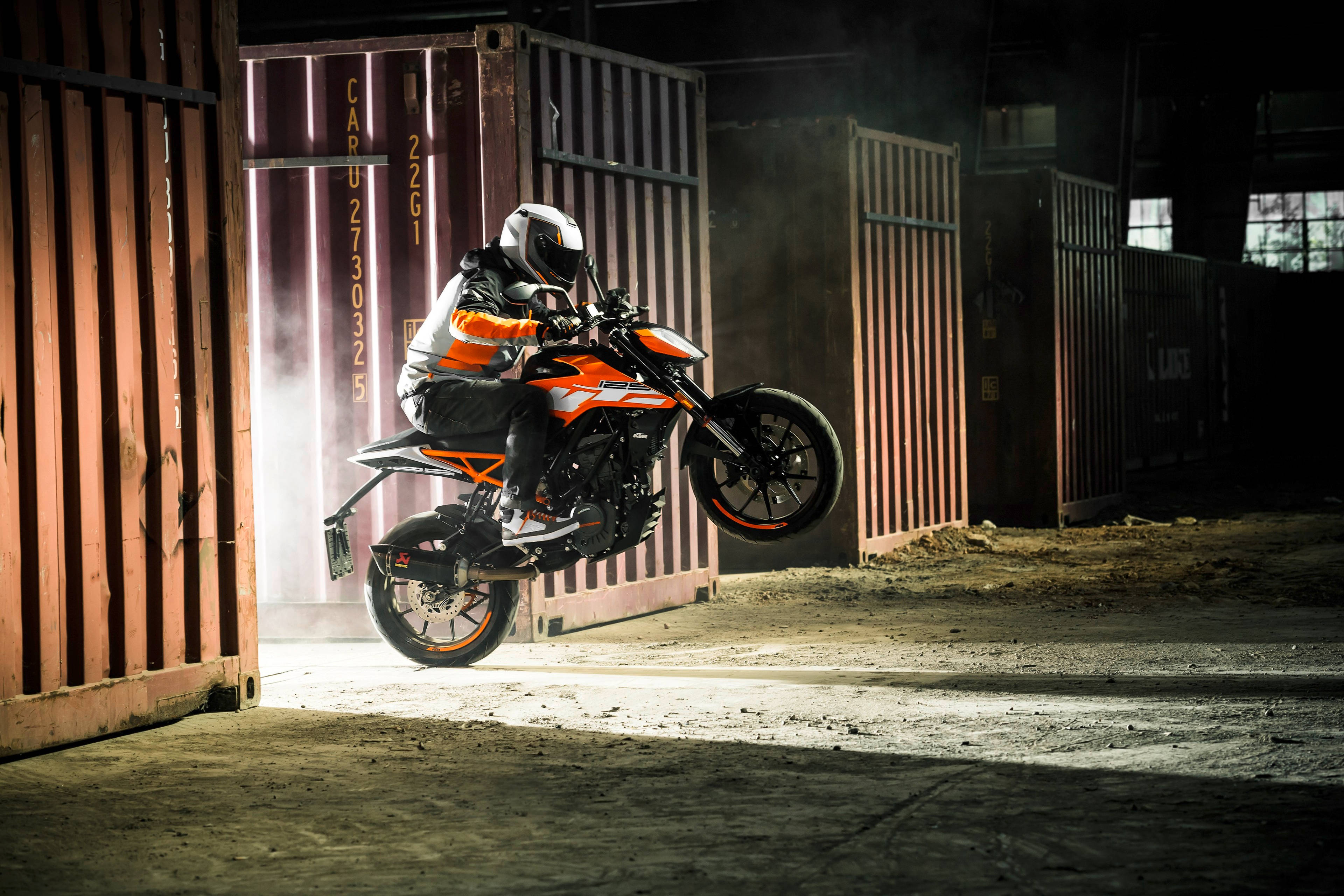 ktm rc 200 桌布 壁紙 — 一名全副裝備的騎手在昏暗的倉庫中，在生鏽的貨運集裝箱之間用橙色KTM電單車表演前輪離地。