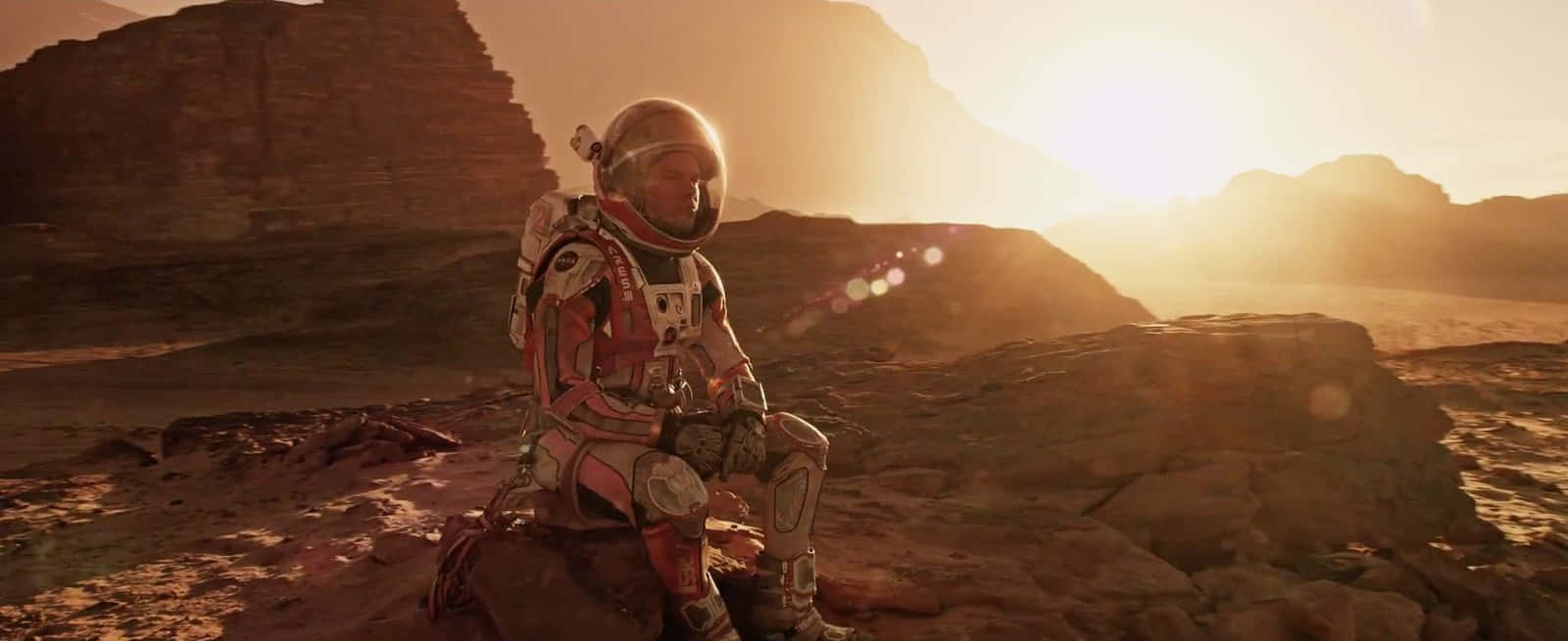 The Martian 桌布 壁紙 — 一名太空人獨自坐在岩石密佈的外星行星上，金色的太陽在遠山後升起，投下長長的影子。