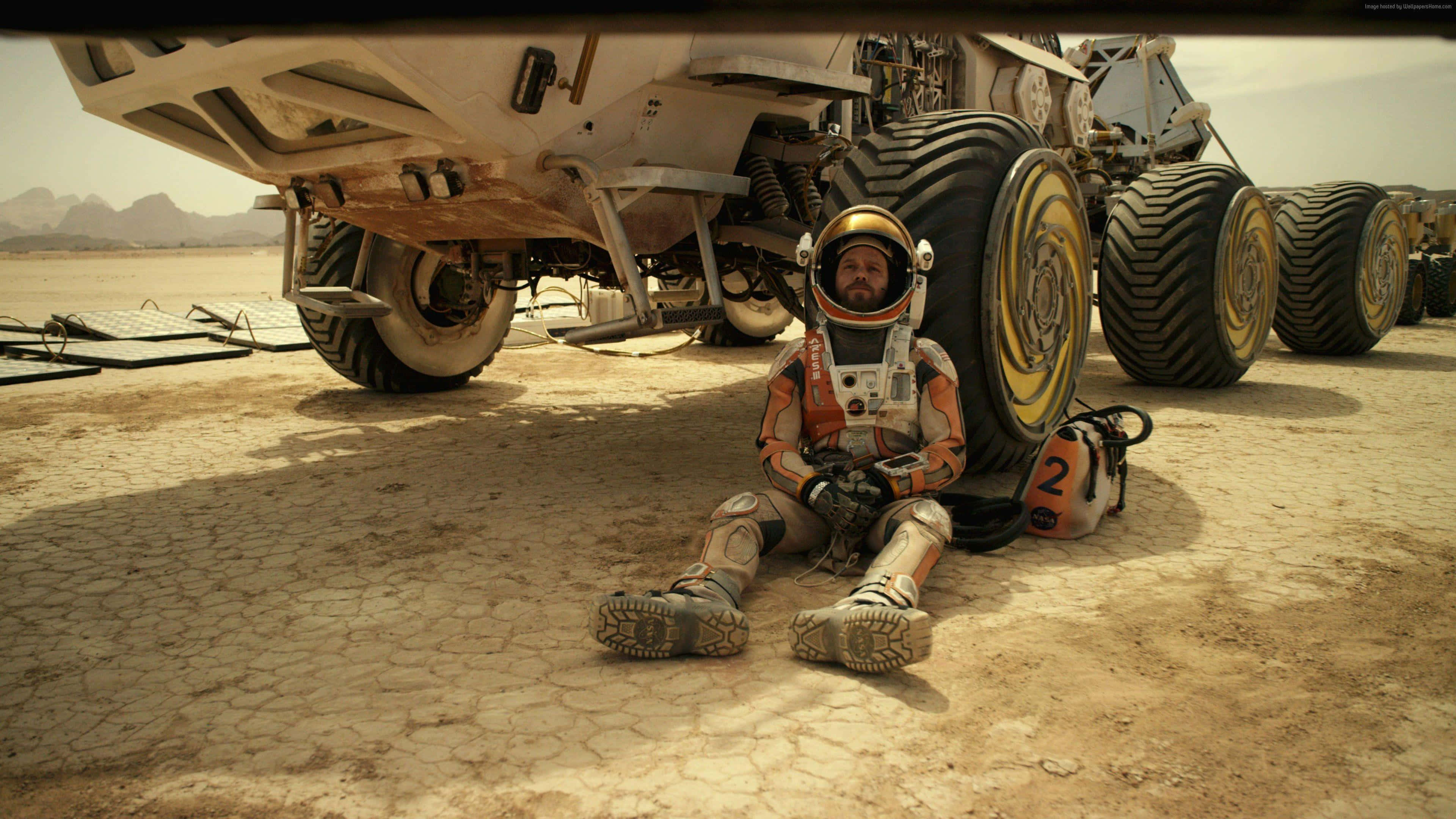 The Martian 桌布 壁紙 — 一名太空人疲憊地坐在佈滿裂縫的火星土壤上，旁邊是一輛巨大的六輪探測車，天空是朦朧的橙色。