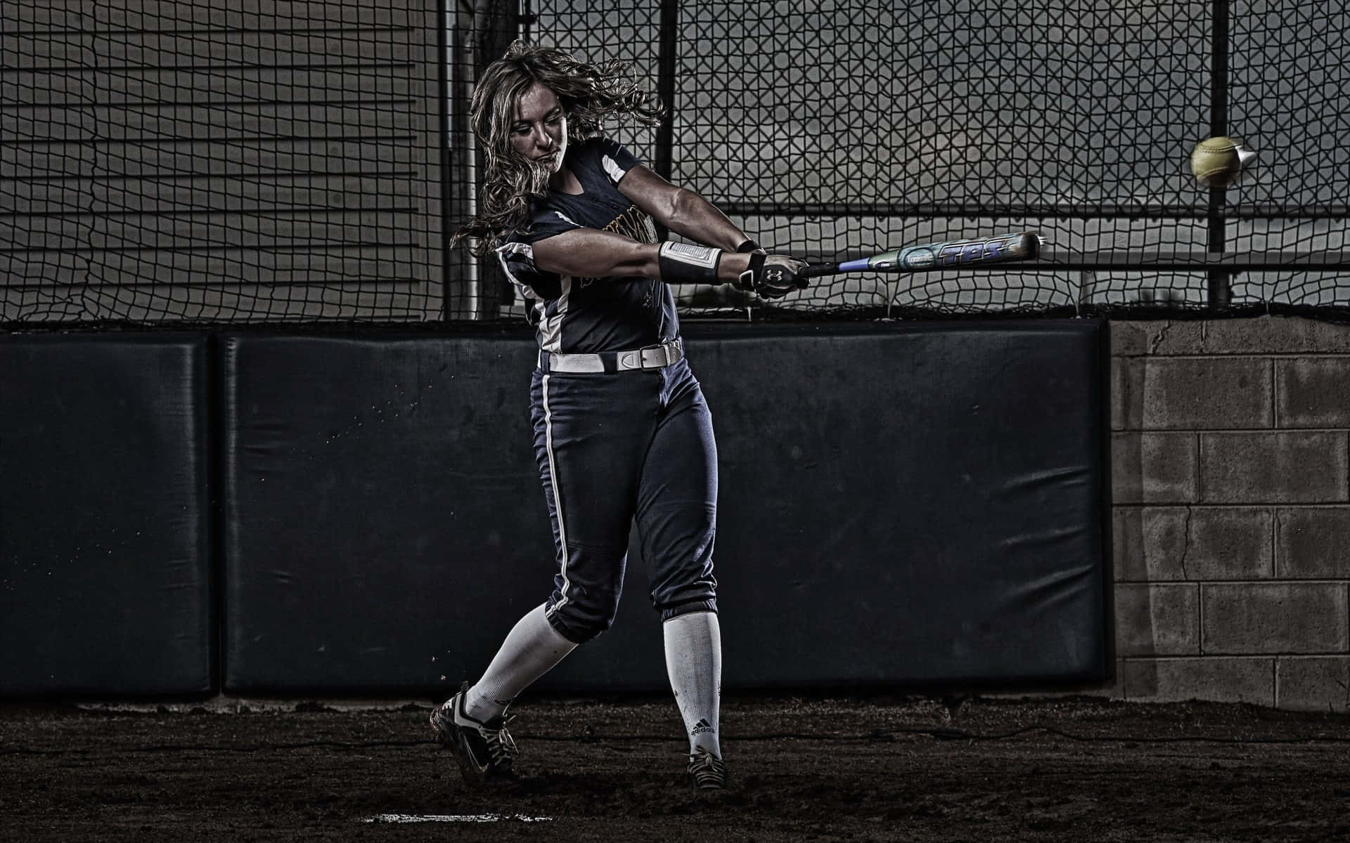 baseball bat 桌布 壁紙 — 一名女壘球運動員在光線昏暗的室內籠中，專注地揮棒，球從旁飛過。