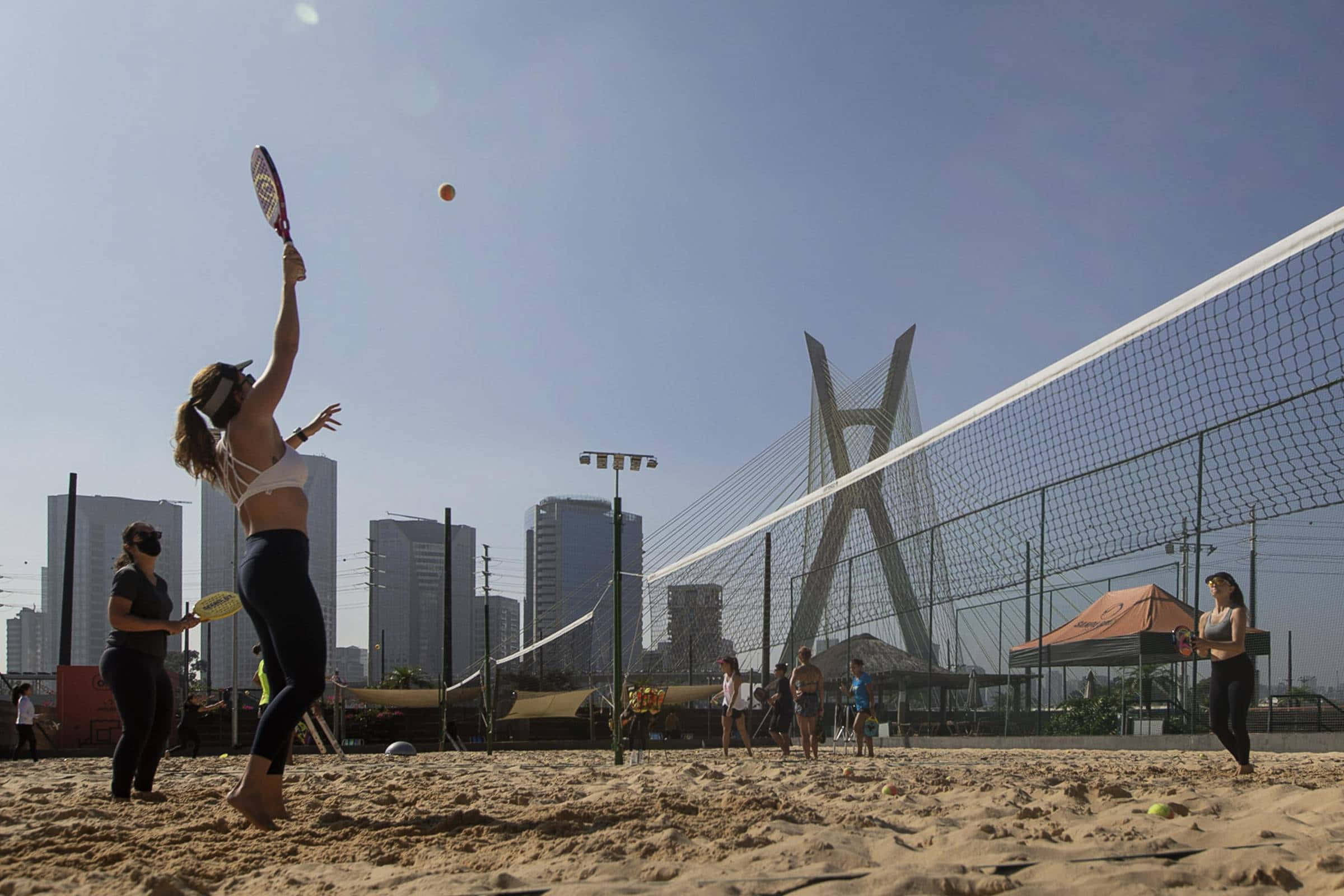 Beach Tennis 桌布 壁紙 — 一名女子在沙灘場地上跳起擊球，背景是現代橋樑和城市天際線，天空湛藍。