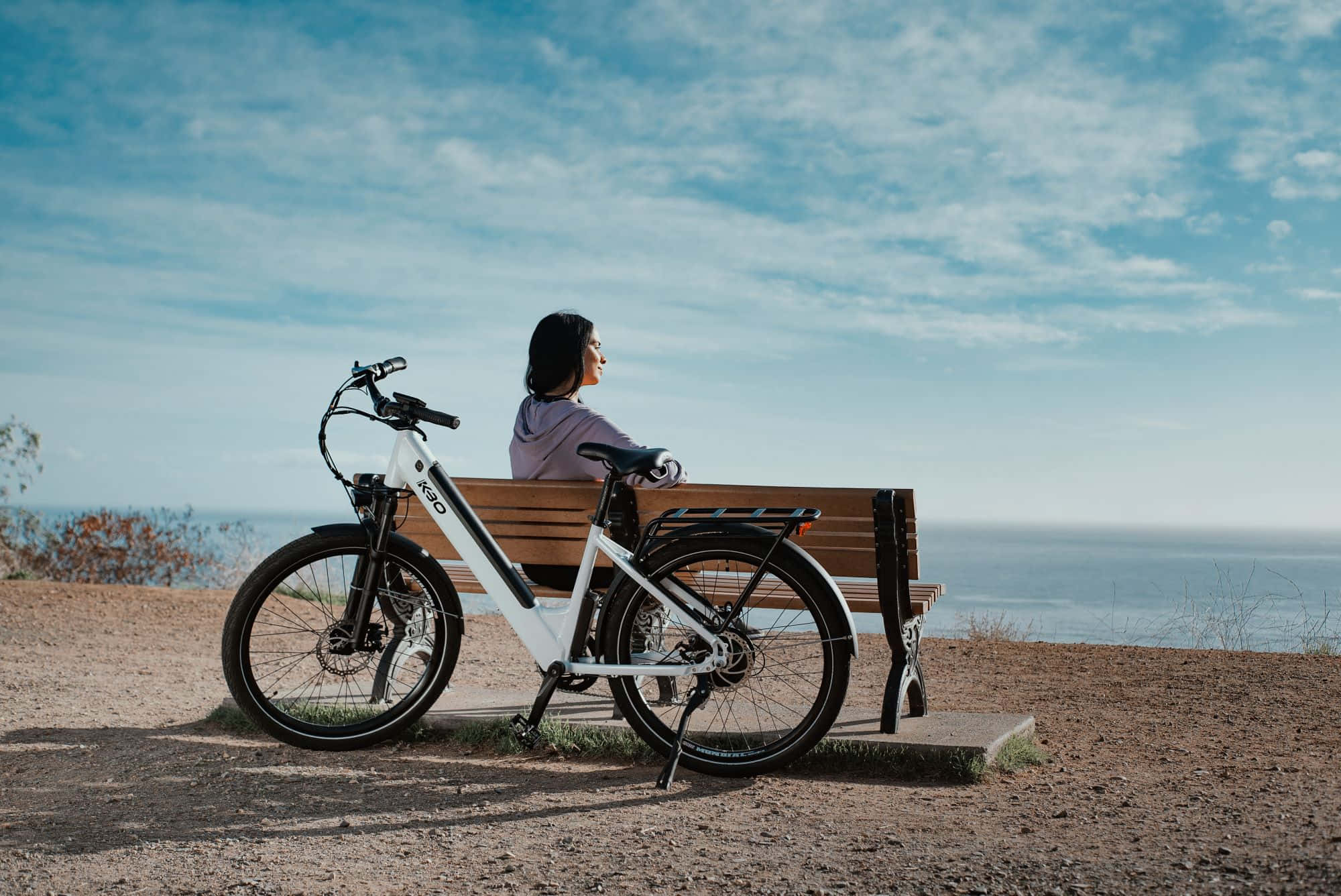Electric Bike 桌布 壁紙 — 一名女子坐在可俯瞰海洋的木長椅上，一輛白色電動單車停在她旁邊，頭頂是明亮的藍天。
