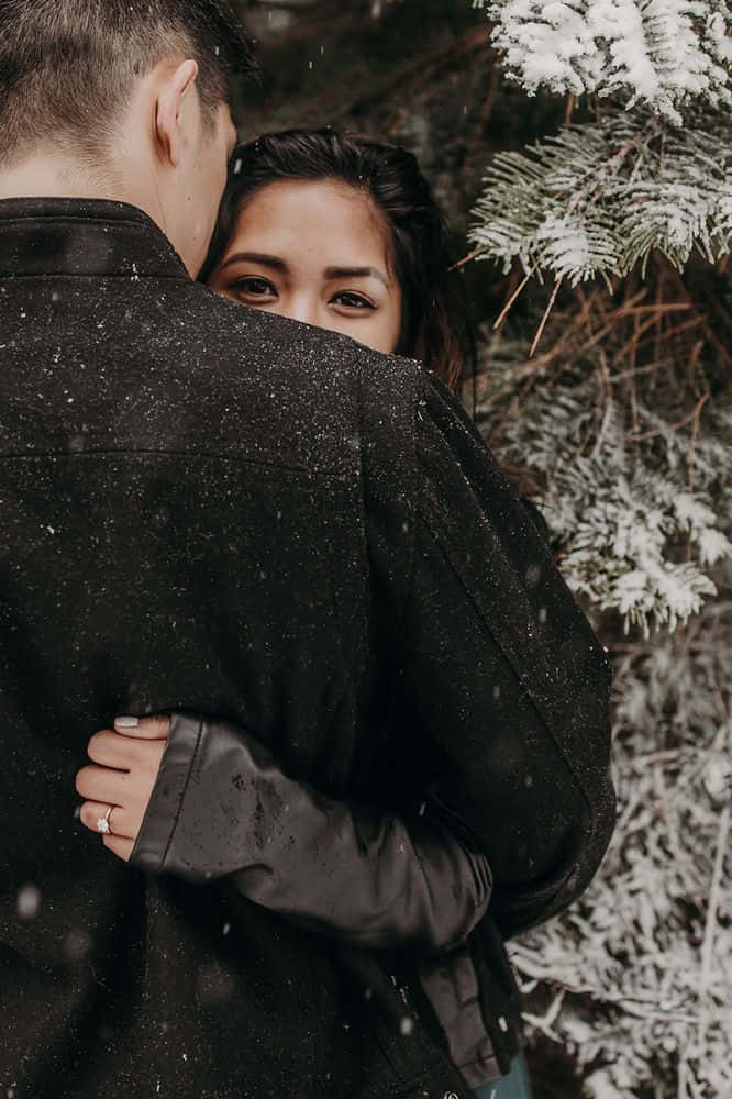 Winter Photography 桌布 壁紙 — 一名女子從一名男子的肩膀後探出頭來，雪花在他們周圍飄落，靠近結霜的松樹枝。