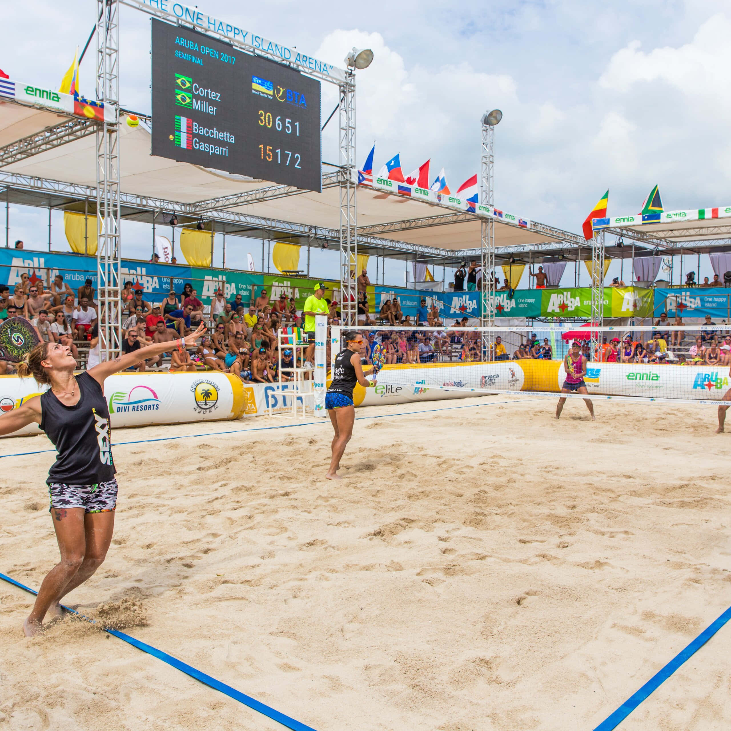 Beach Tennis 桌布 壁紙 — 一名女子沙灘排球運動員在陽光明媚的日間比賽中跳起扣球，背景有觀眾和記分牌。