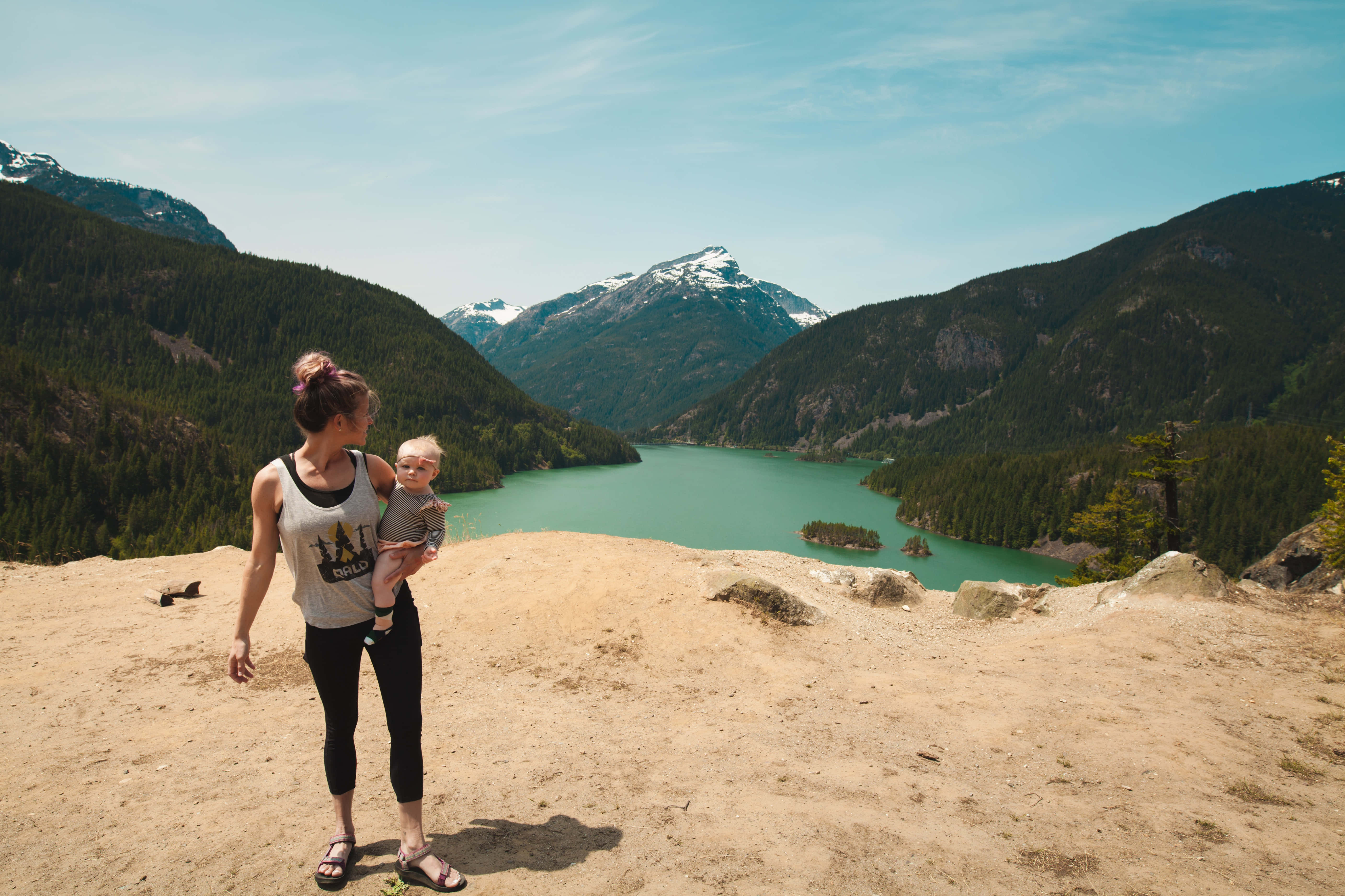 Mom 桌布 壁紙 — 一名女子站在陽光普照的懸崖上，懷裡抱著一個嬰兒，俯瞰著一個被森林覆蓋的山脈和積雪山峰環抱的碧綠湖泊。