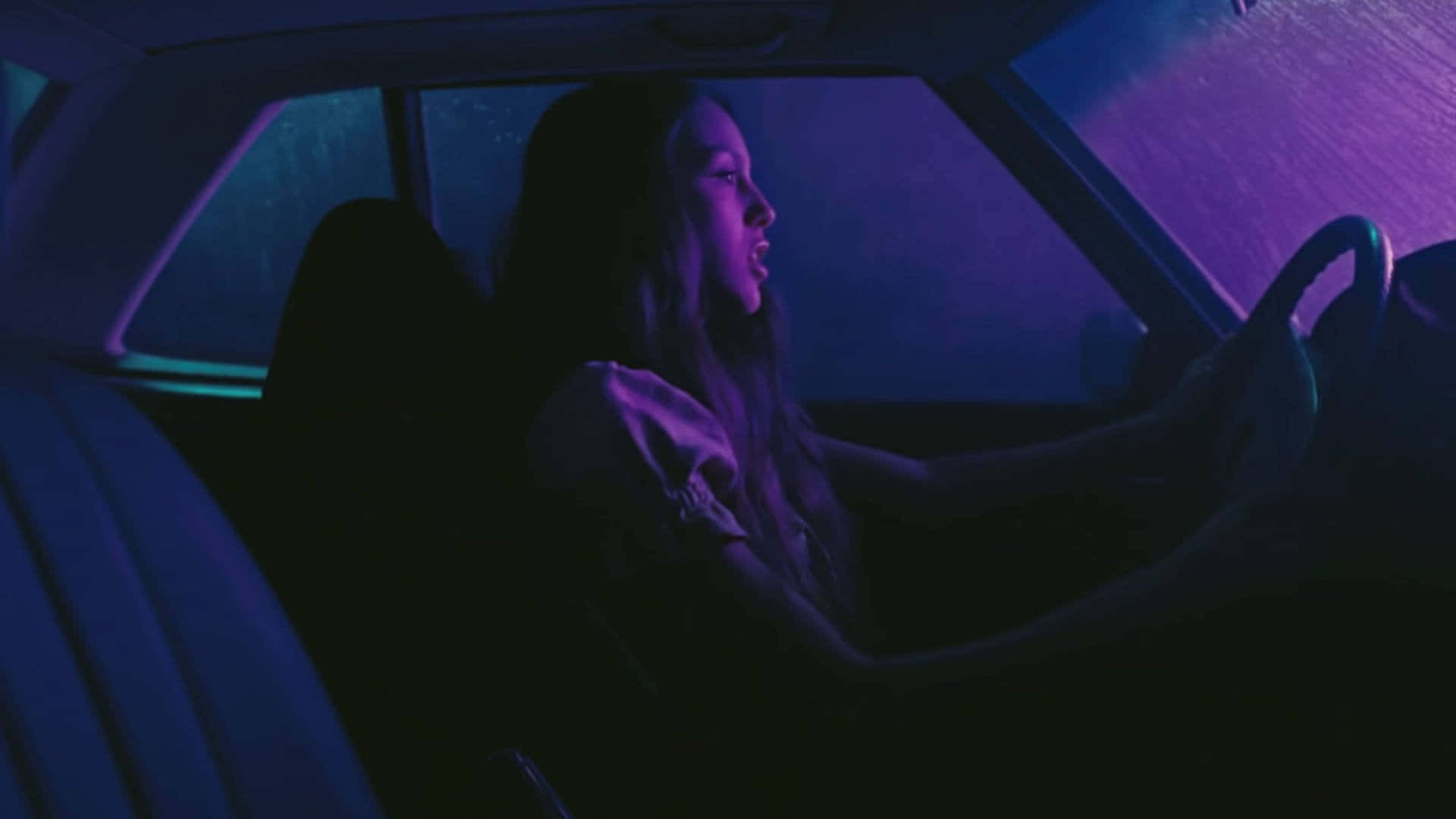 Olivia Rodrigo 桌布 壁紙 — 一名女性在夜間駕駛汽車，車內被街道標誌的霓虹紫色和藍色燈光籠罩。