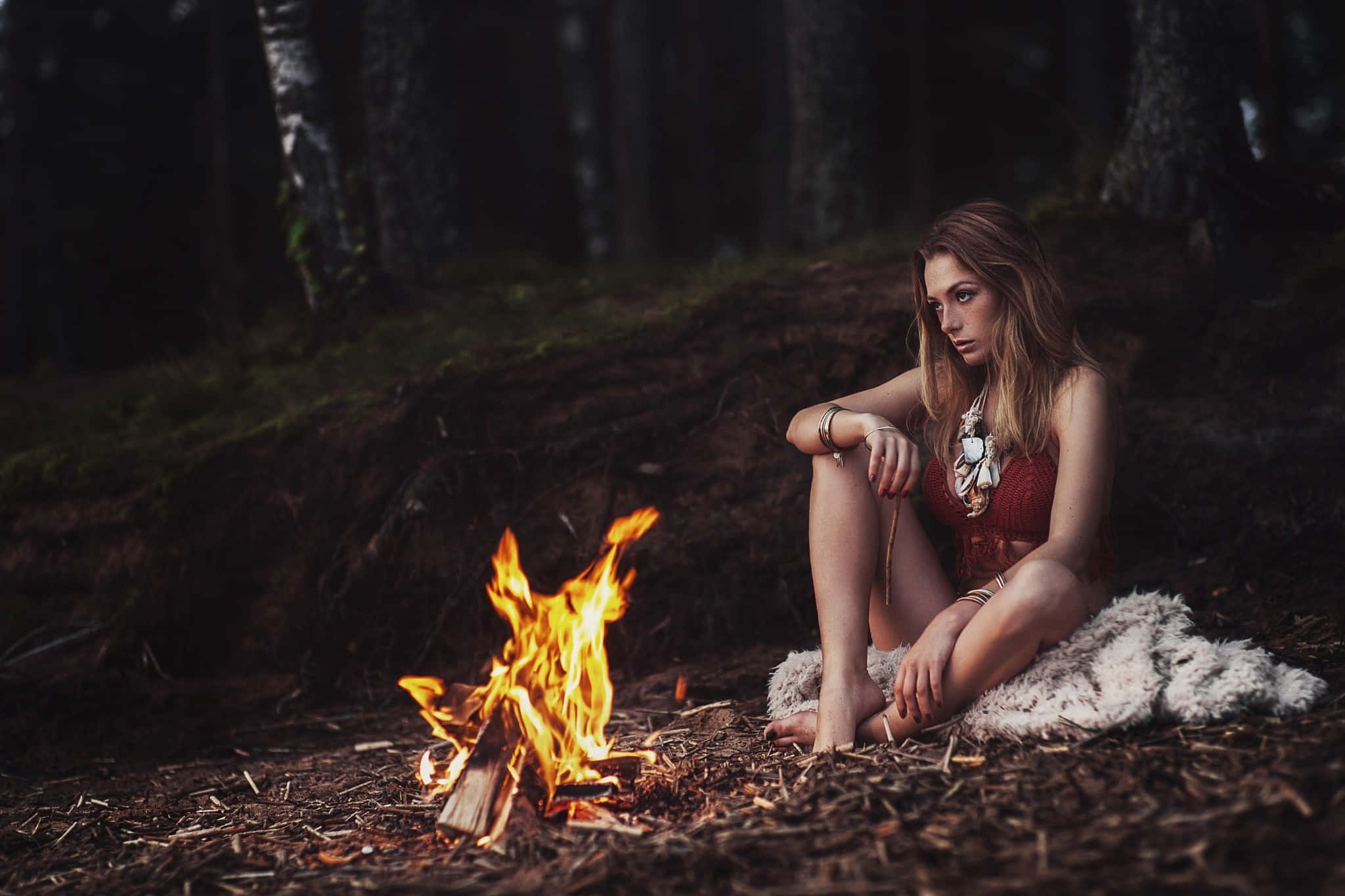 Fall Bonfire 桌布 壁紙 — 一名女性赤腳坐在白色毛皮地毯上，旁邊是黑暗森林中噼啪作響的營火，在夜晚。