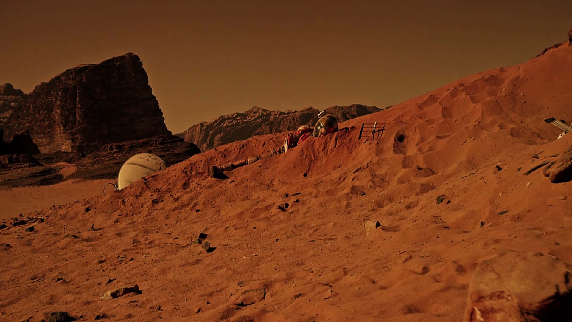 The Martian 桌布 壁紙 — 一名孤獨的太空人在朦朧的橙色天空下探索崎嶇的紅色火星景觀，附近有岩石露頭和圓頂棲息地。
