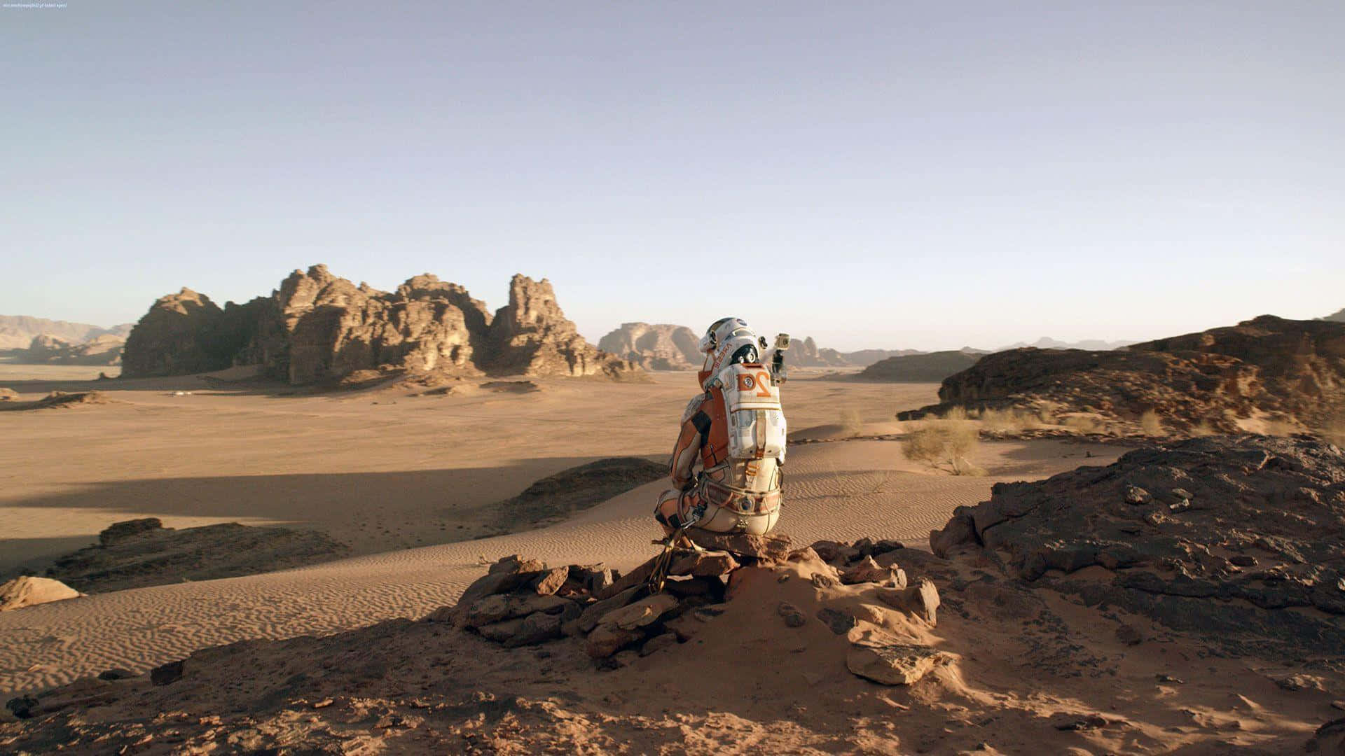 The Martian 桌布 壁紙 — 一名孤獨的太空人穿著白色太空衣，跪在岩石沙漠山脊上，俯瞰廣闊的沙丘和遠處的岩石地貌，天空晴朗。