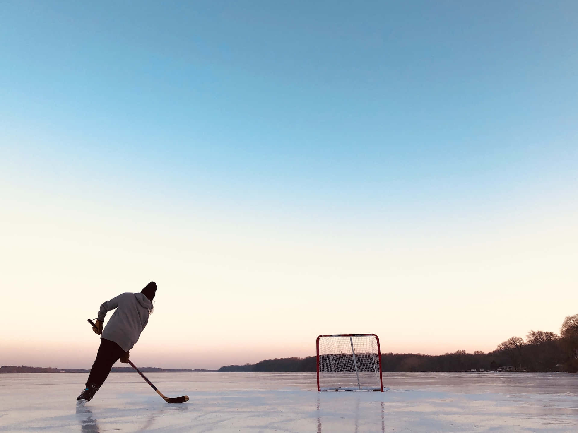 Ice Rink 桌布 壁紙 — 一名孤獨的曲棍球運動員在日落時分，在結冰的湖面上，在清晰的漸變天空下滑向球門。