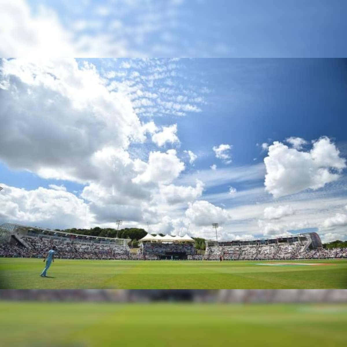 Cricket Ground 桌布 壁紙 — 一名孤獨的板球運動員在廣闊的藍天下，點綴著蓬鬆的白雲，走過充滿活力的綠色球場。