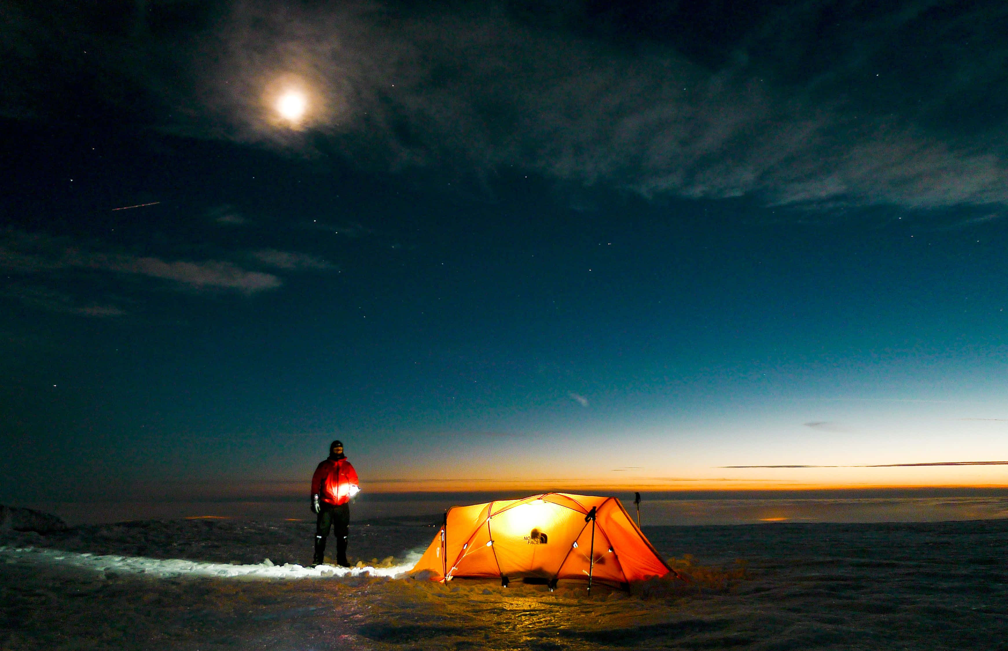 Beach Camping 桌布 壁紙 — 一名孤獨的露營者站在發光的橙色帳篷旁，在明亮的月亮和星空下，雪原之上。