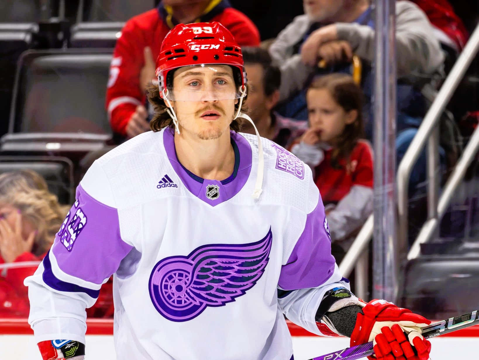 Tyler Bertuzzi 桌布 壁紙 — 一名專注的曲棍球運動員身穿白色和紫色球衣，帶有翼輪標誌，站在冰上手持球棍。
