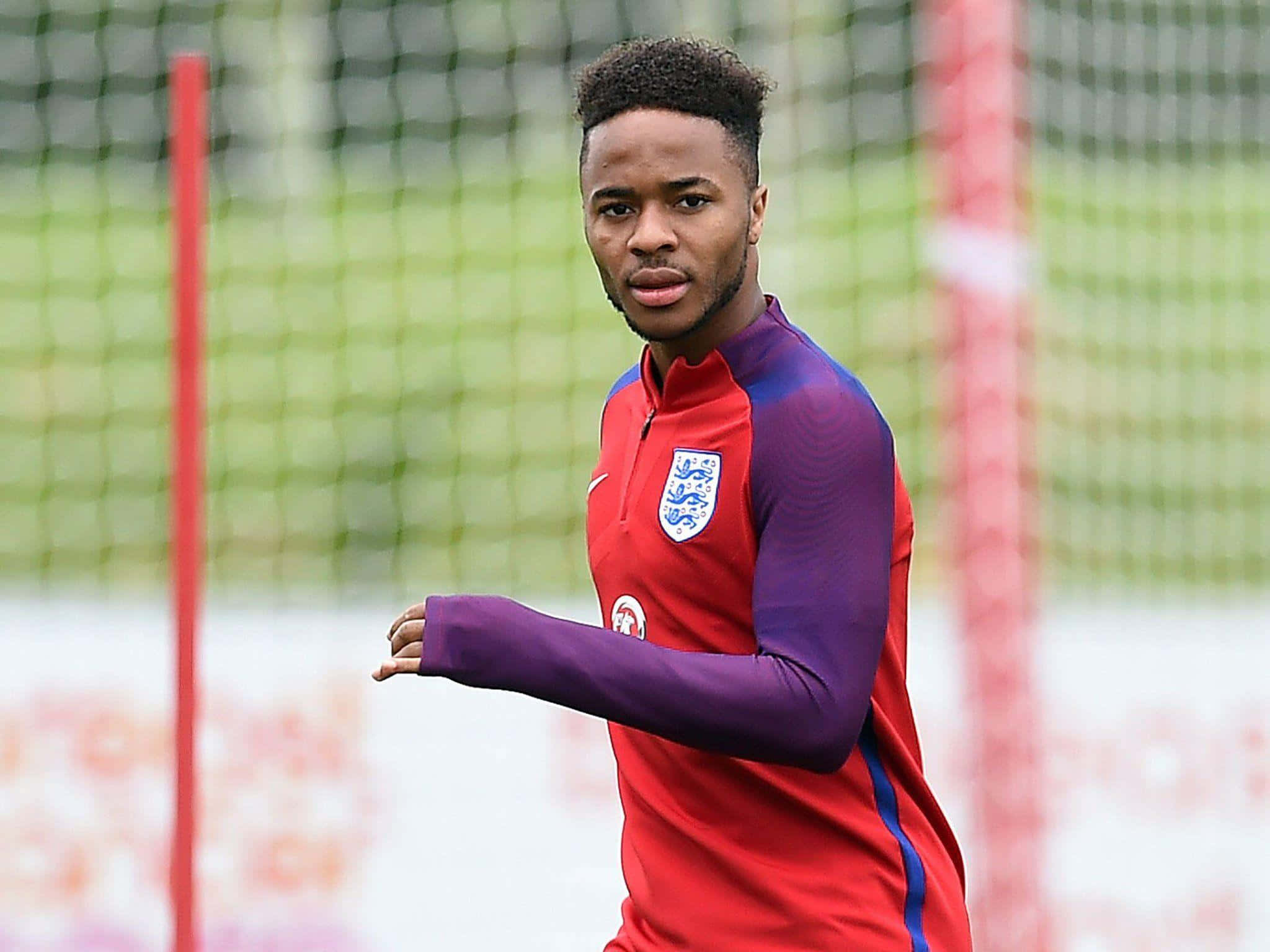 Sterling 桌布 壁紙 — 一名專注的英格蘭足球員身穿紅色和紫色訓練裝備，在草地上向前邁步，身後是模糊的球門網。