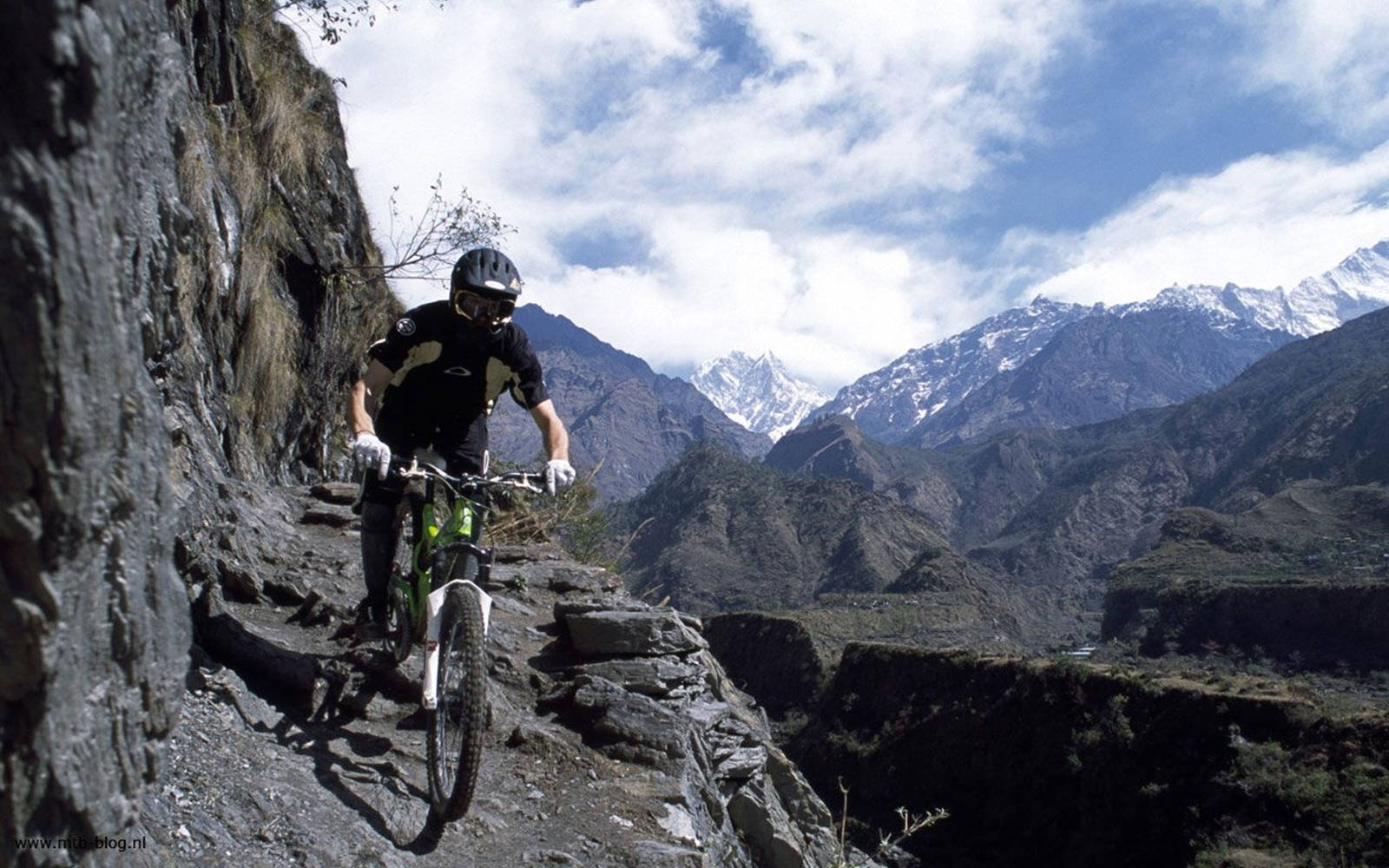 Mtb 桌布 壁紙 — 一名山地單車手在部分多雲的天空下，沿著陡峭懸崖旁的狹窄岩石小徑行駛，背景中聳立著白雪覆蓋的山峰。